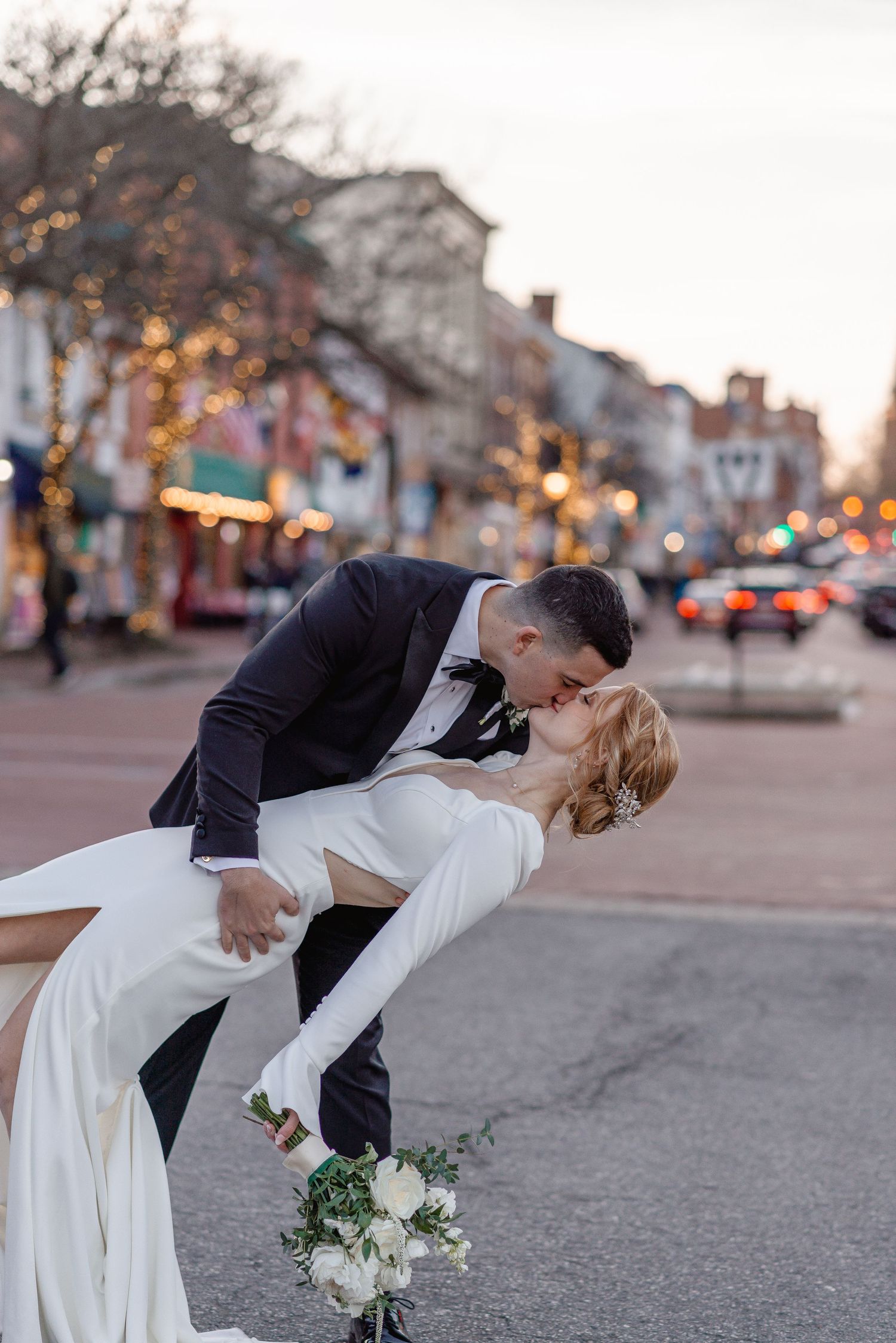 Bride and groom share romantic dip kiss on Main Street Annapolis at dusk with bokeh city lights.