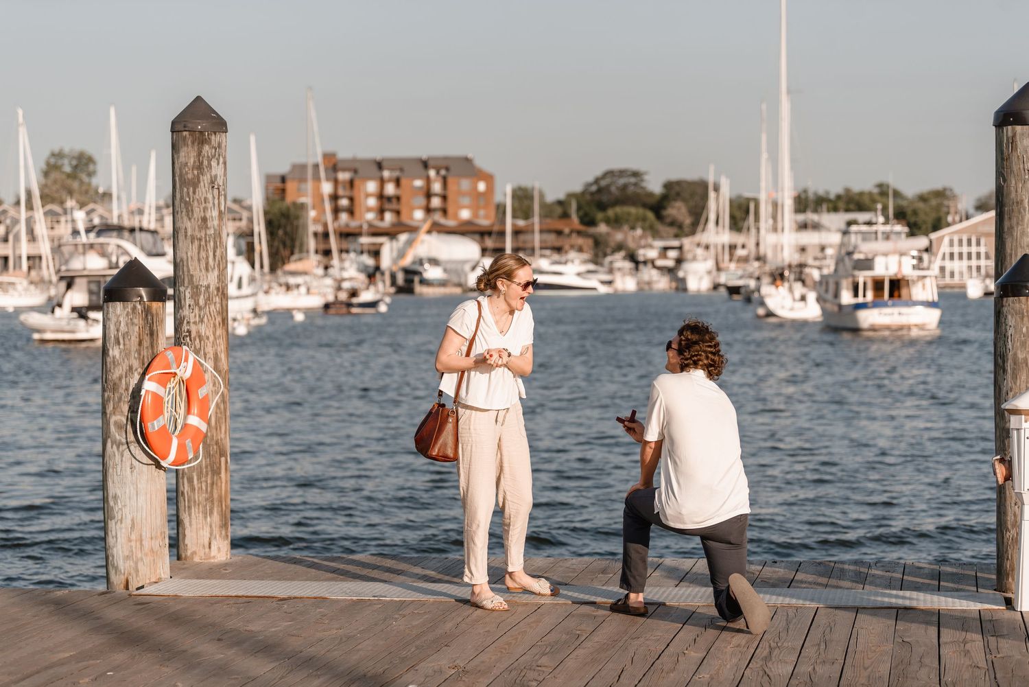Man on one knee proposing to his girlfriend on a scenic waterfront dock at golden hour.