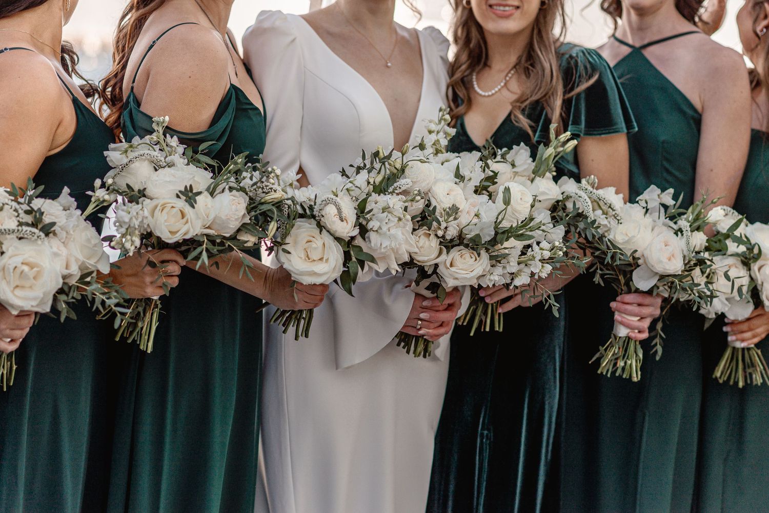 Bride in white gown with bridesmaids in emerald green dresses holding white rose and peony bouquets.