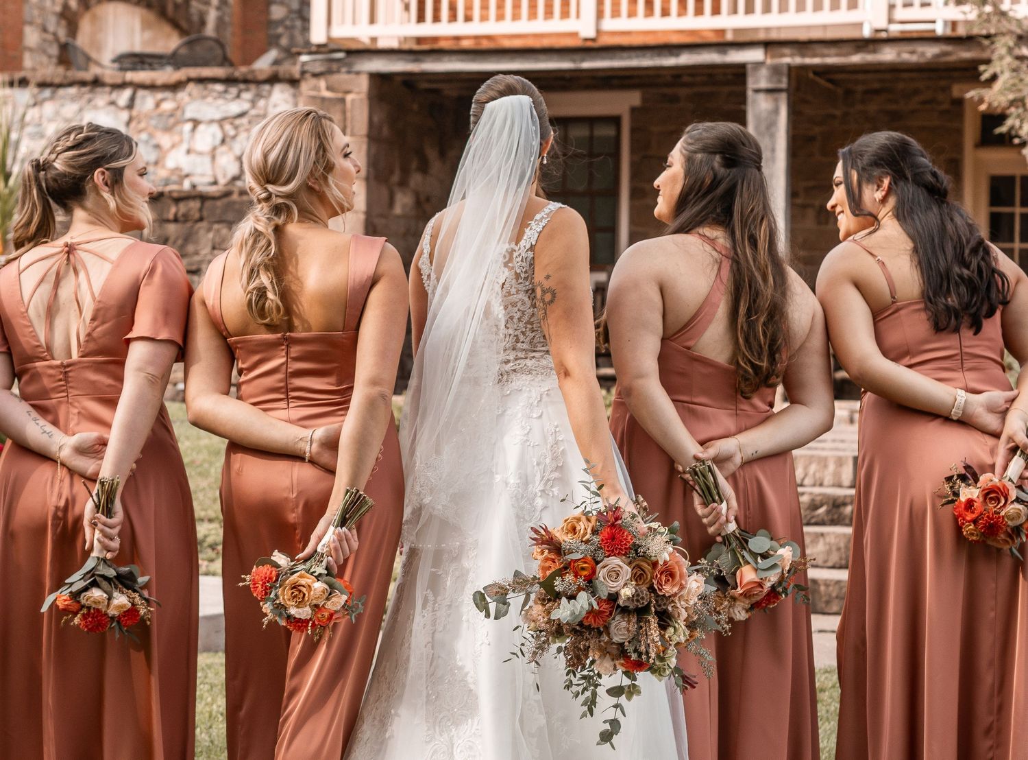 Bride and bridesmaids in rust orange dresses holding fall bouquets, viewed from behind at outdoor wedding.