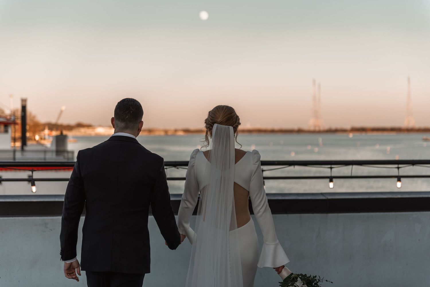 Bride in backless gown and groom hold hands gazing at a moonrise over the water at sunset.