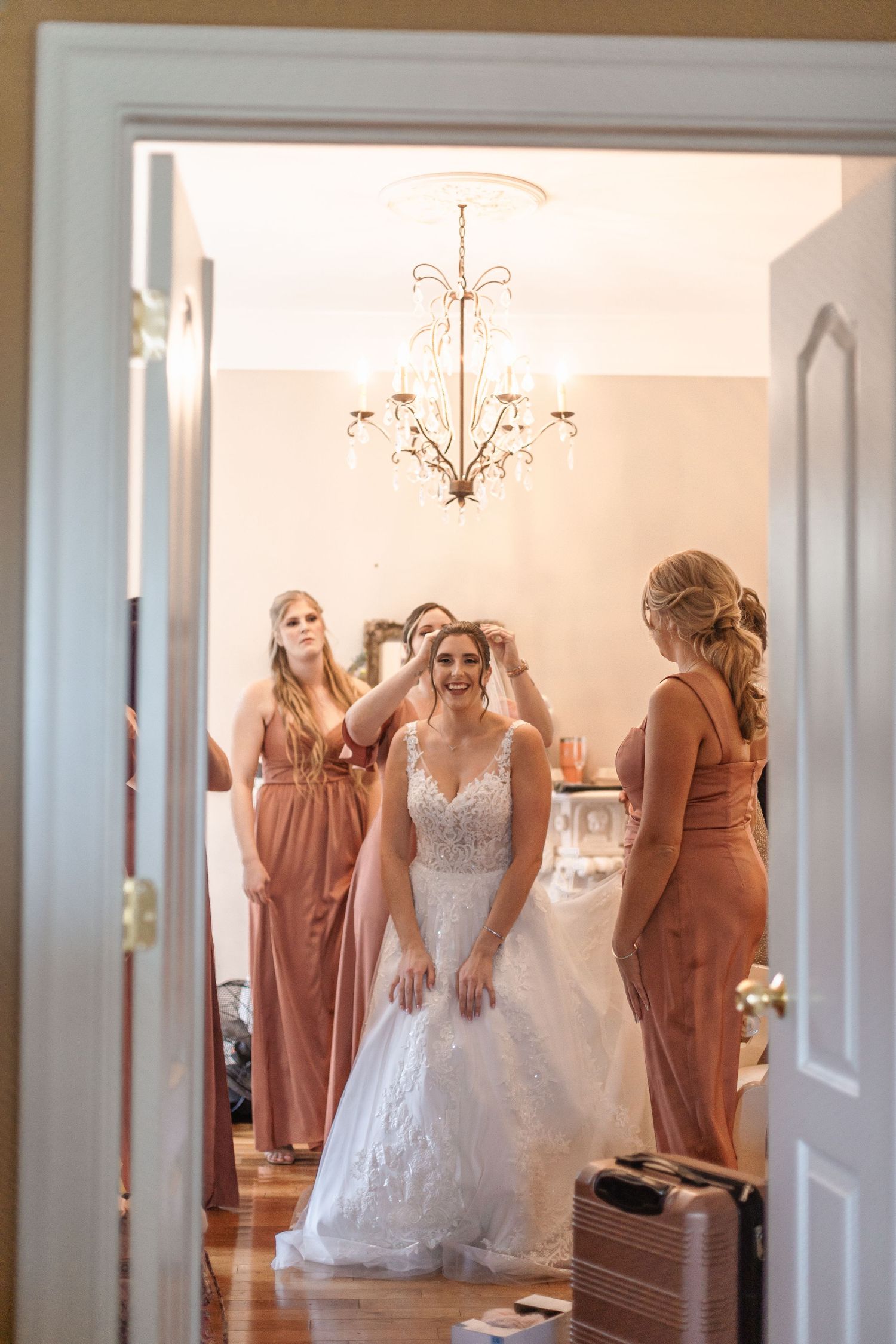 Bridesmaids in dusty rose dresses help the smiling bride with her veil in a warmly lit bridal suite.