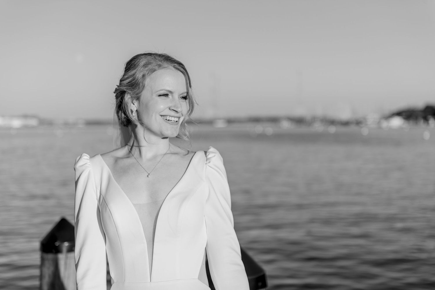 Black and white portrait of a radiant bride in a deep-V white gown smiling by the waterfront.