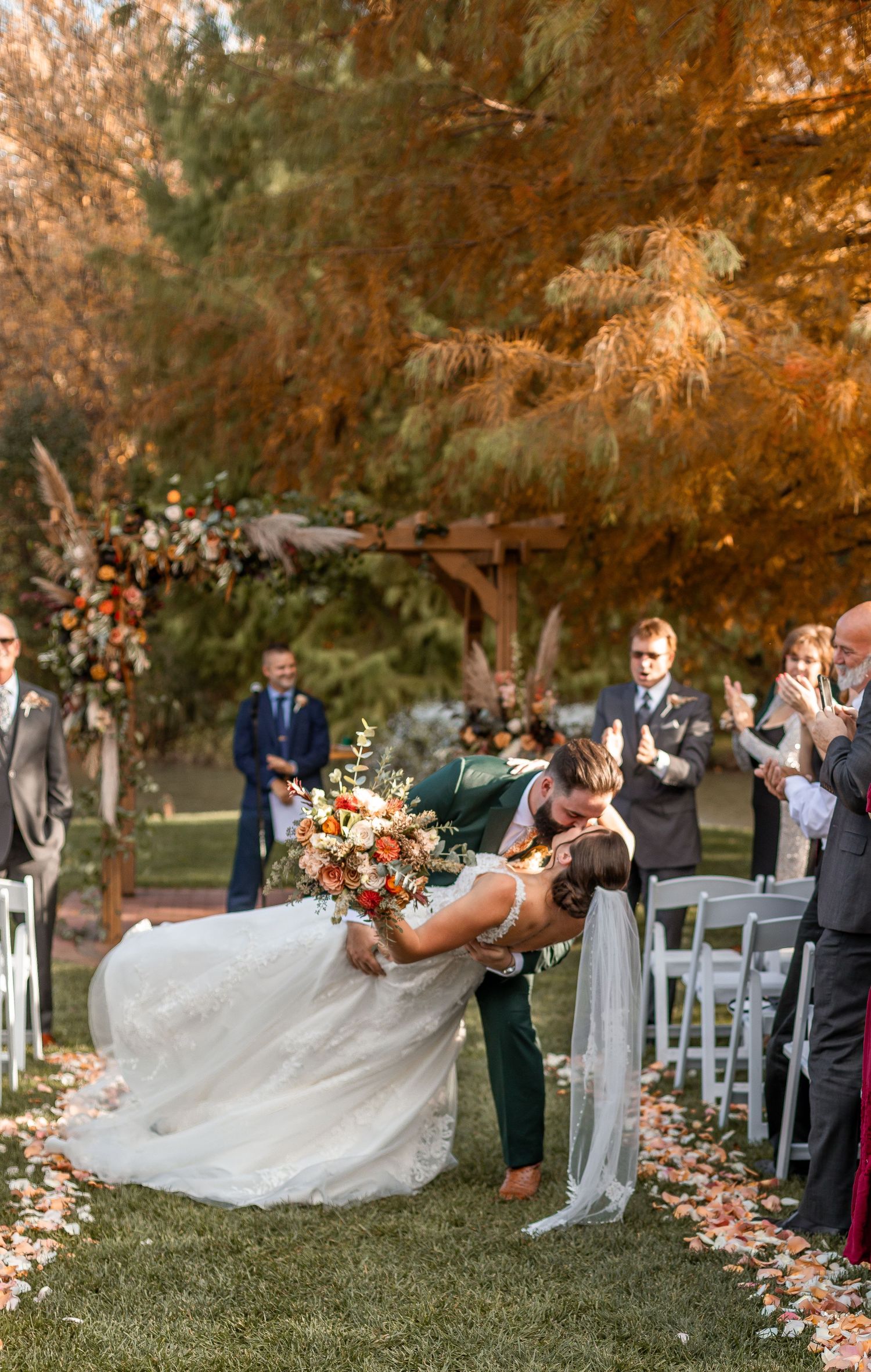 Groom dips bride for a passionate kiss while guests applaud along a fall foliage-lined outdoor wedding ceremony aisle.