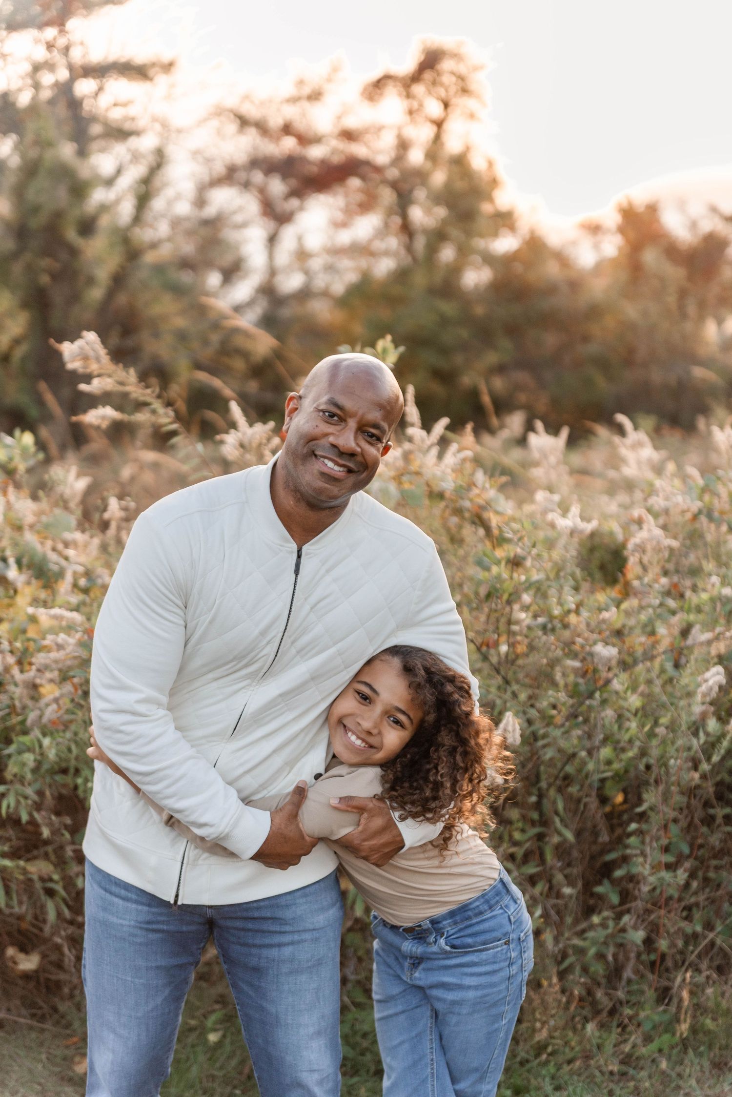 A smiling bald father hugs his curly-haired child in a sunlit field with golden hour lighting.