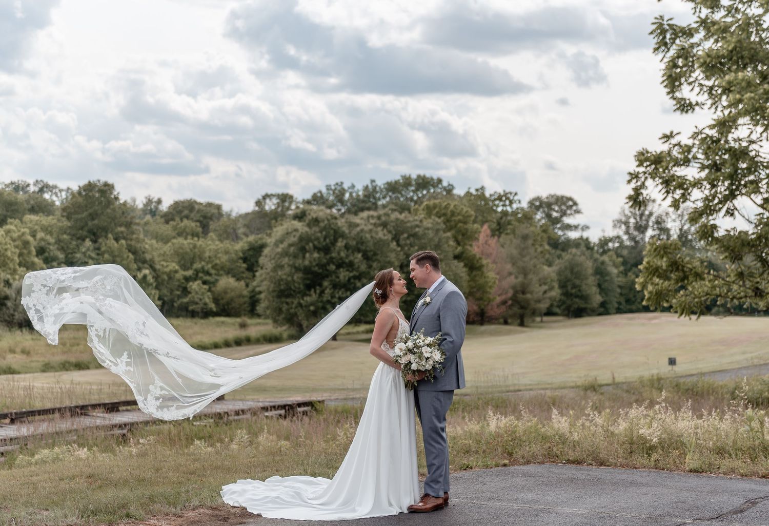 Bride and groom share romantic moment outdoors as long lace veil flows dramatically in the wind.