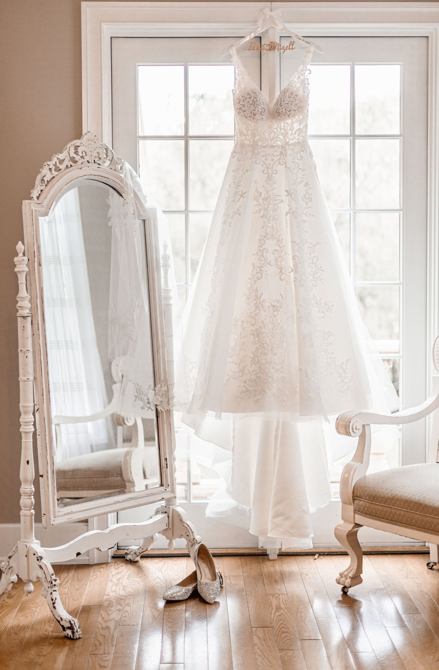 Elegant white lace A-line wedding dress hanging by a window beside an ornate vintage mirror and chair.
