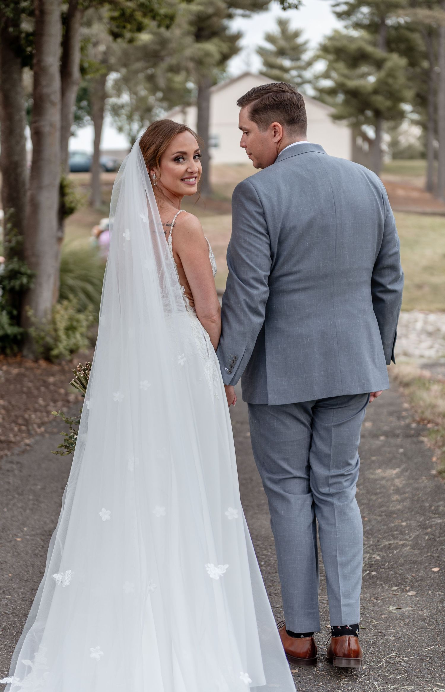 Bride in flowing veil and groom in gray suit walk hand-in-hand along a tree-lined path.