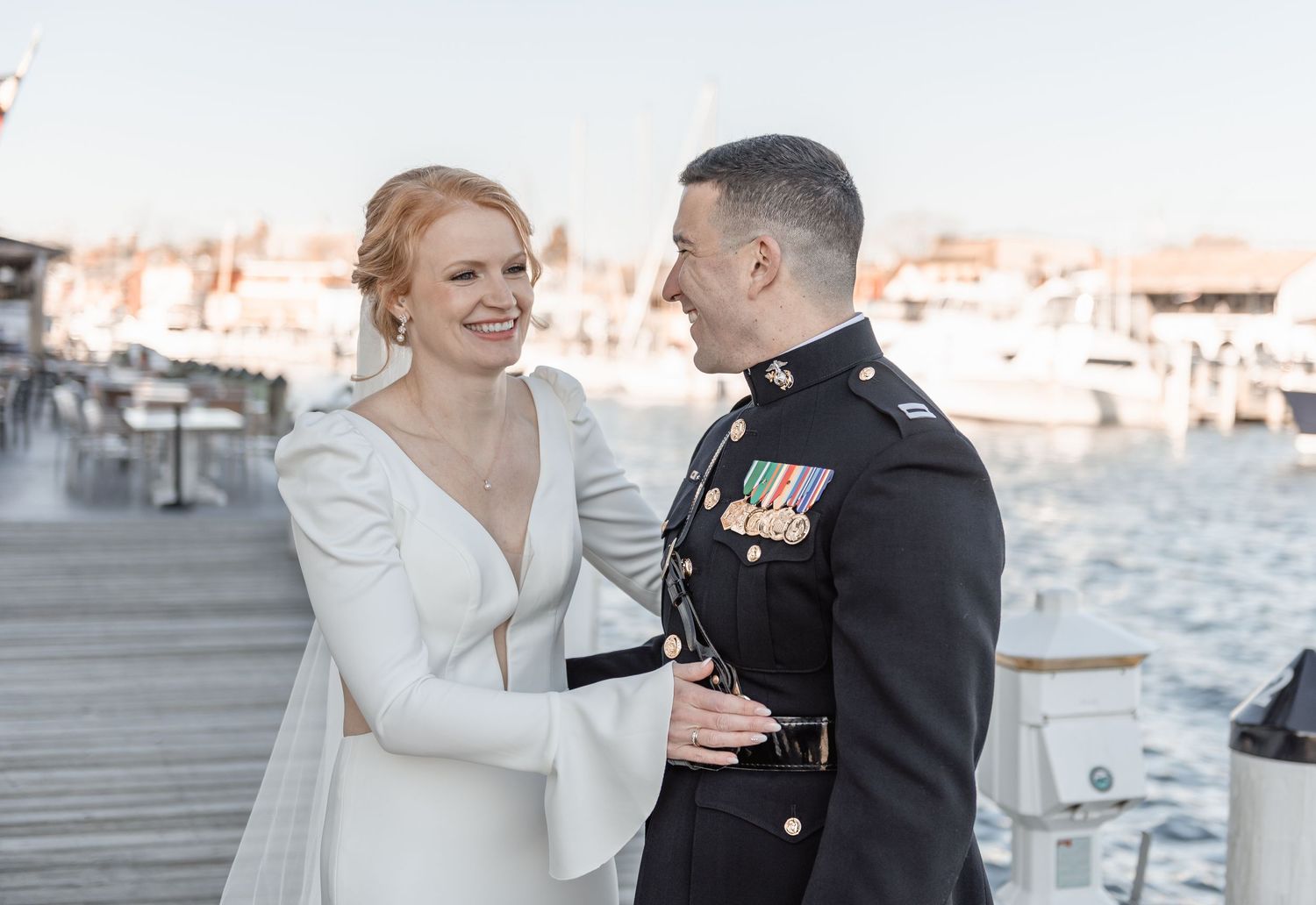 Bride in white gown and groom in military dress uniform share a joyful moment on a waterfront dock.