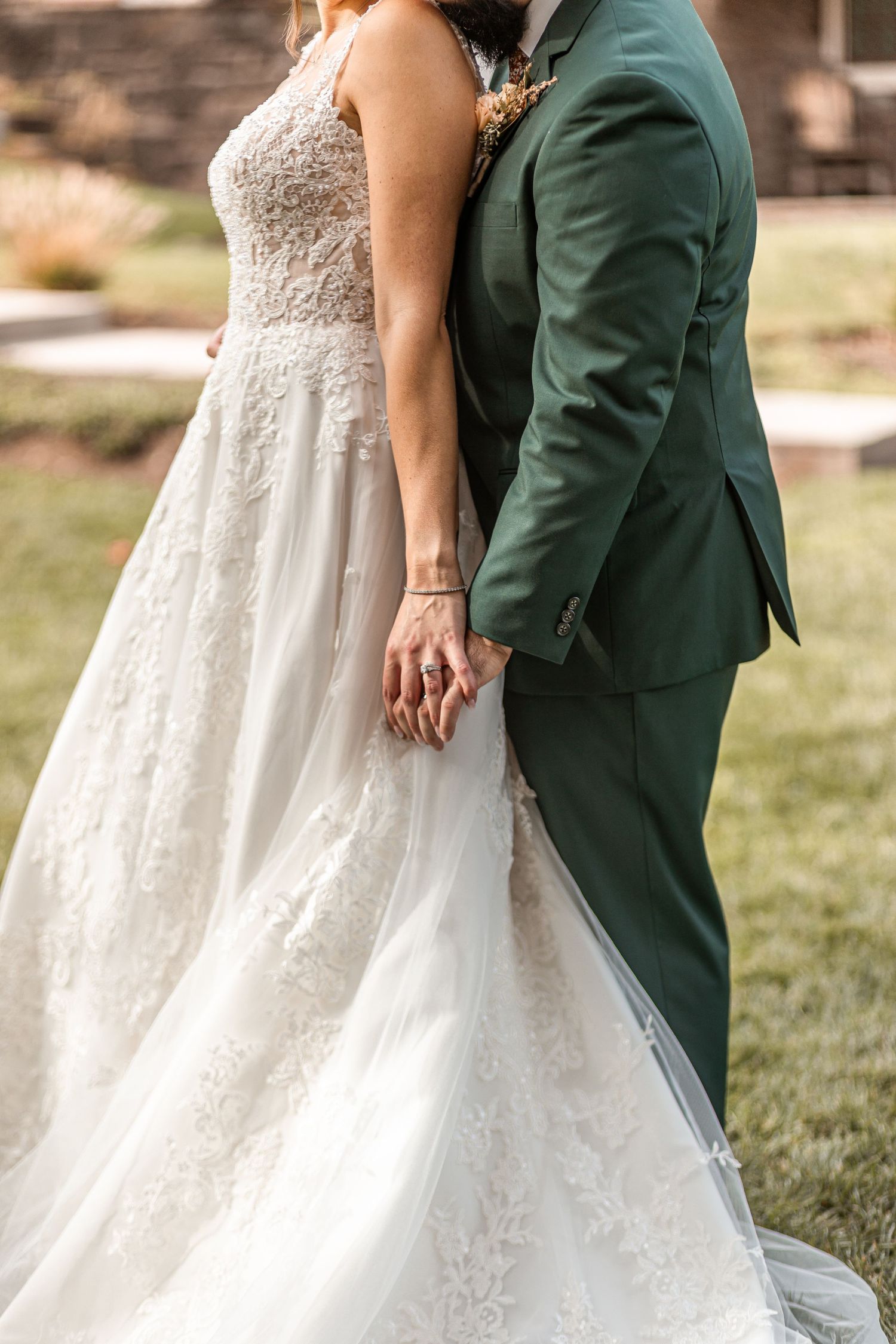 Bride and groom holding hands, she in lace wedding gown, he in dark green suit, standing together outdoors.