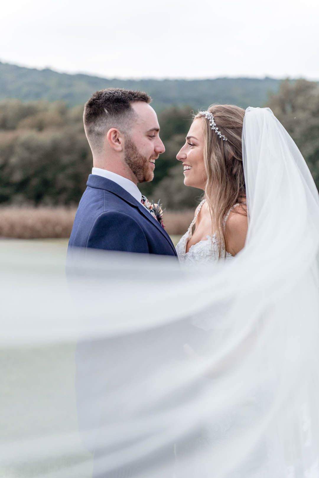Smiling bride and groom embrace outdoors with flowing veil and scenic lake backdrop on wedding day.