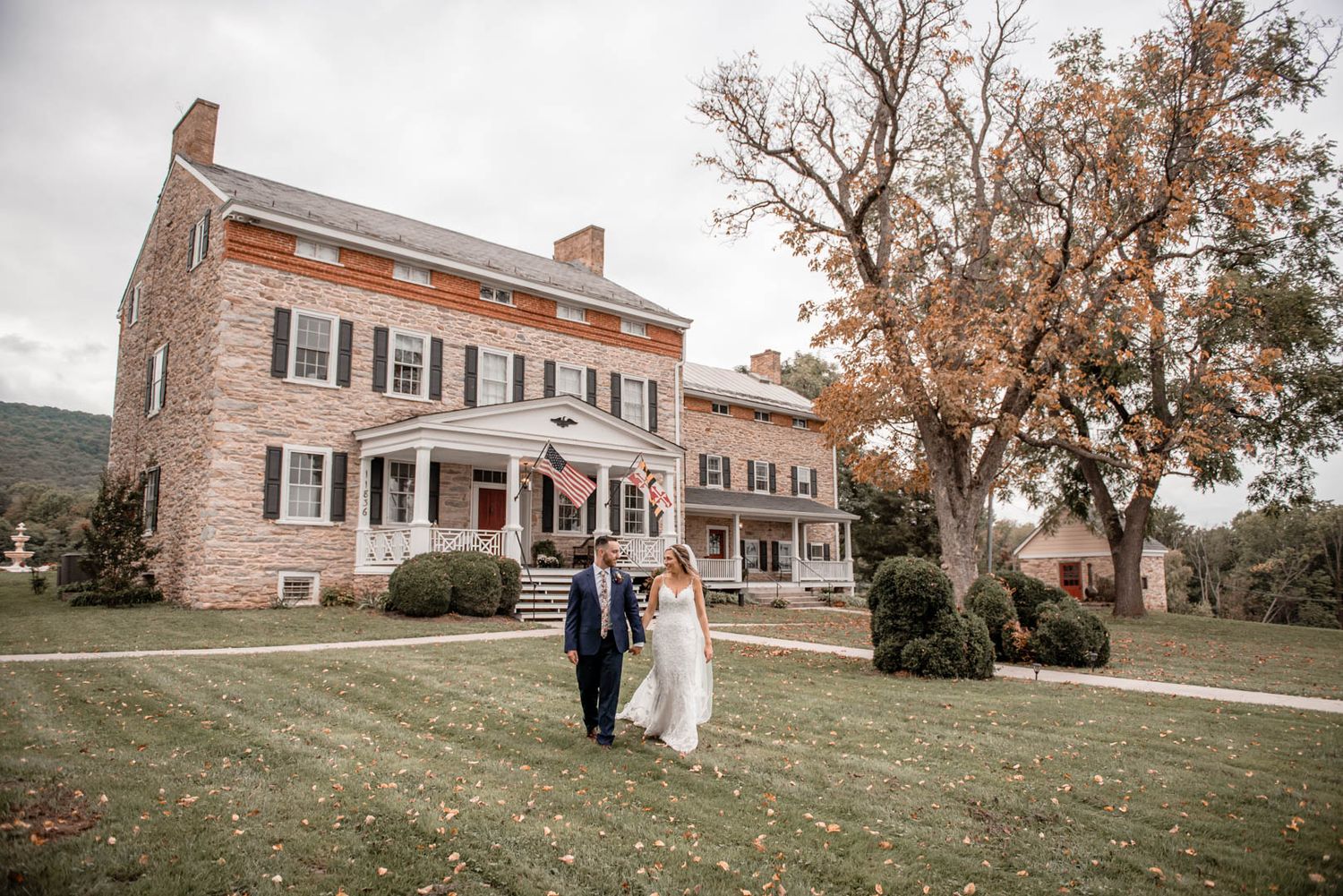 Bride and groom walk hand in hand across a lush lawn in front of an elegant historic brick estate.