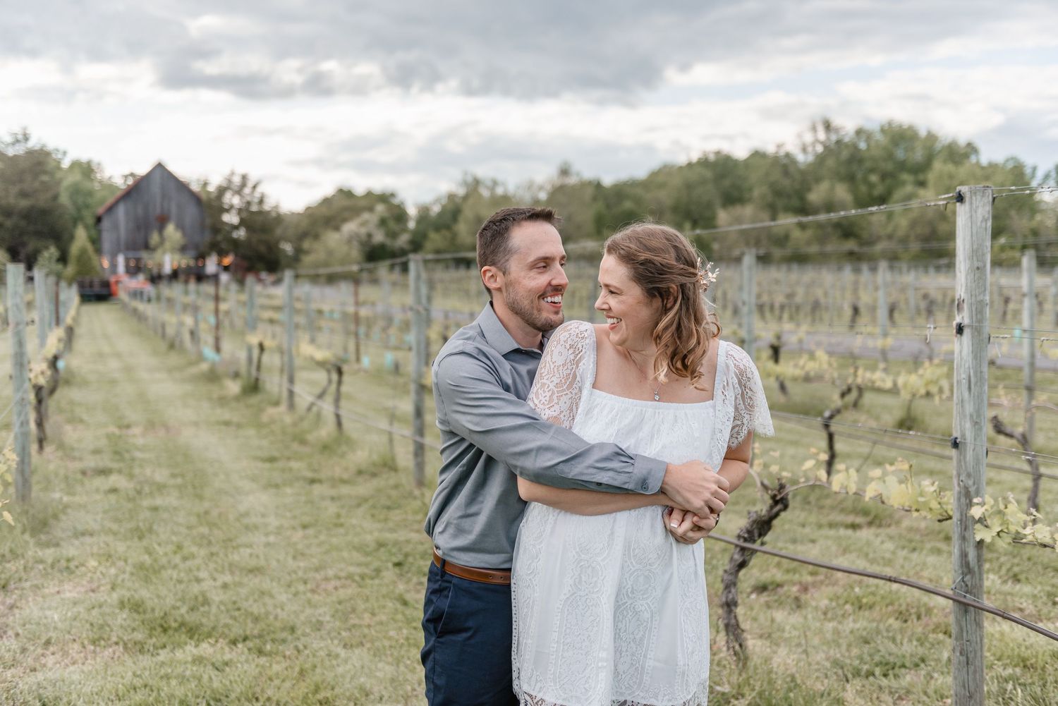 Couple laughing and embracing along a vineyard fence row with a rustic barn in the background.