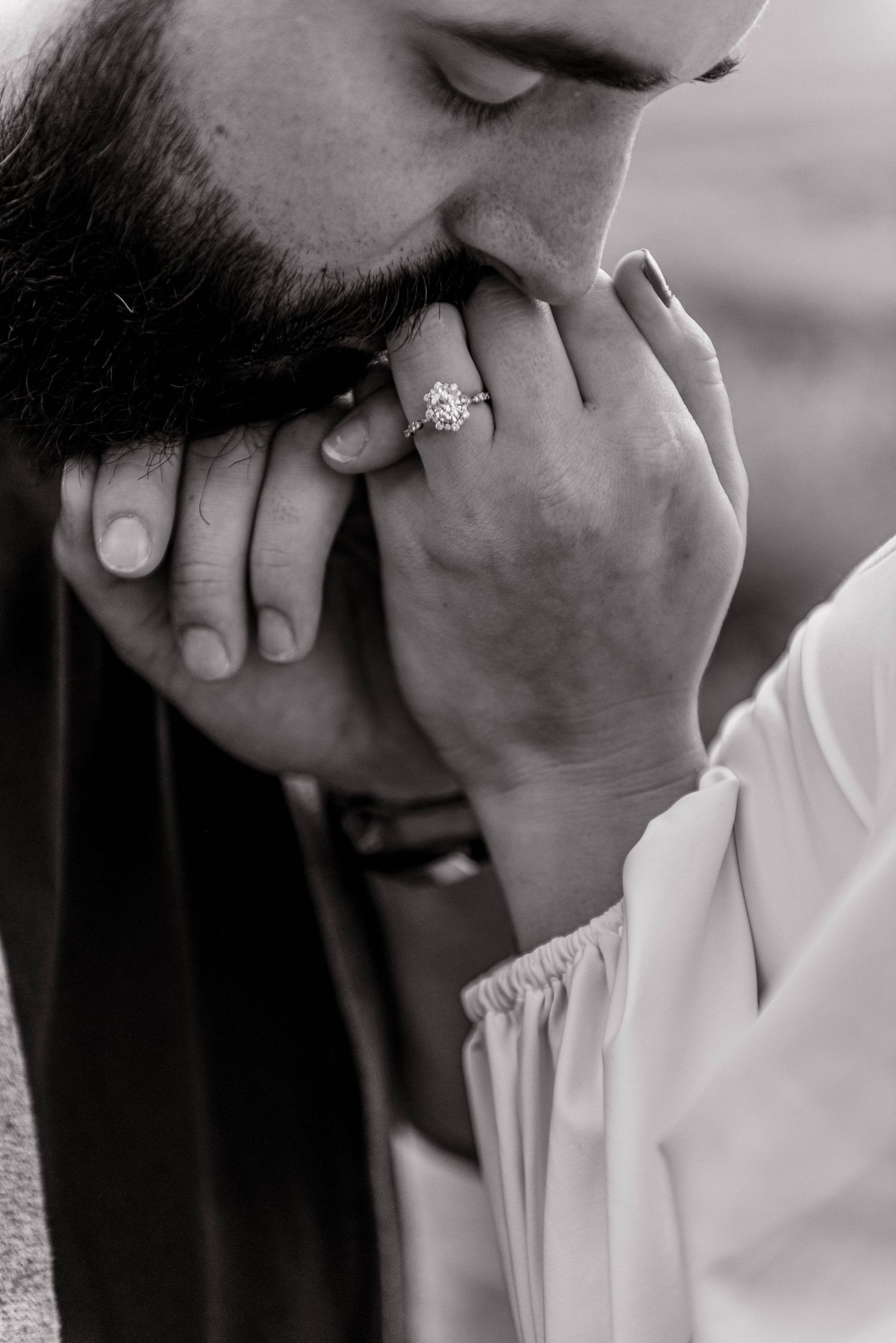 Close-up black and white photo of a man kissing a woman's hand adorned with a diamond engagement ring.