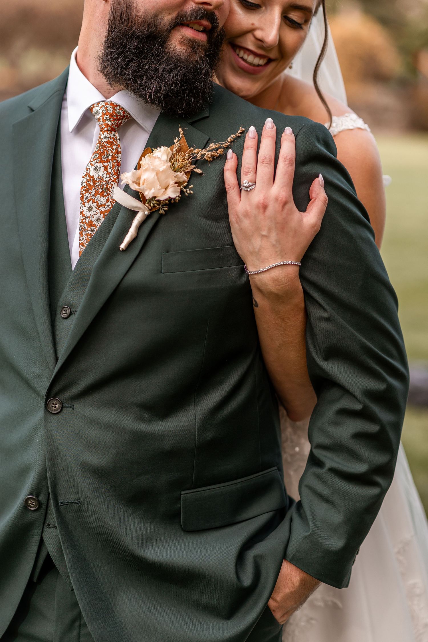 Close-up of bride resting hand on groom's dark green suit with floral boutonniere, showing ring.