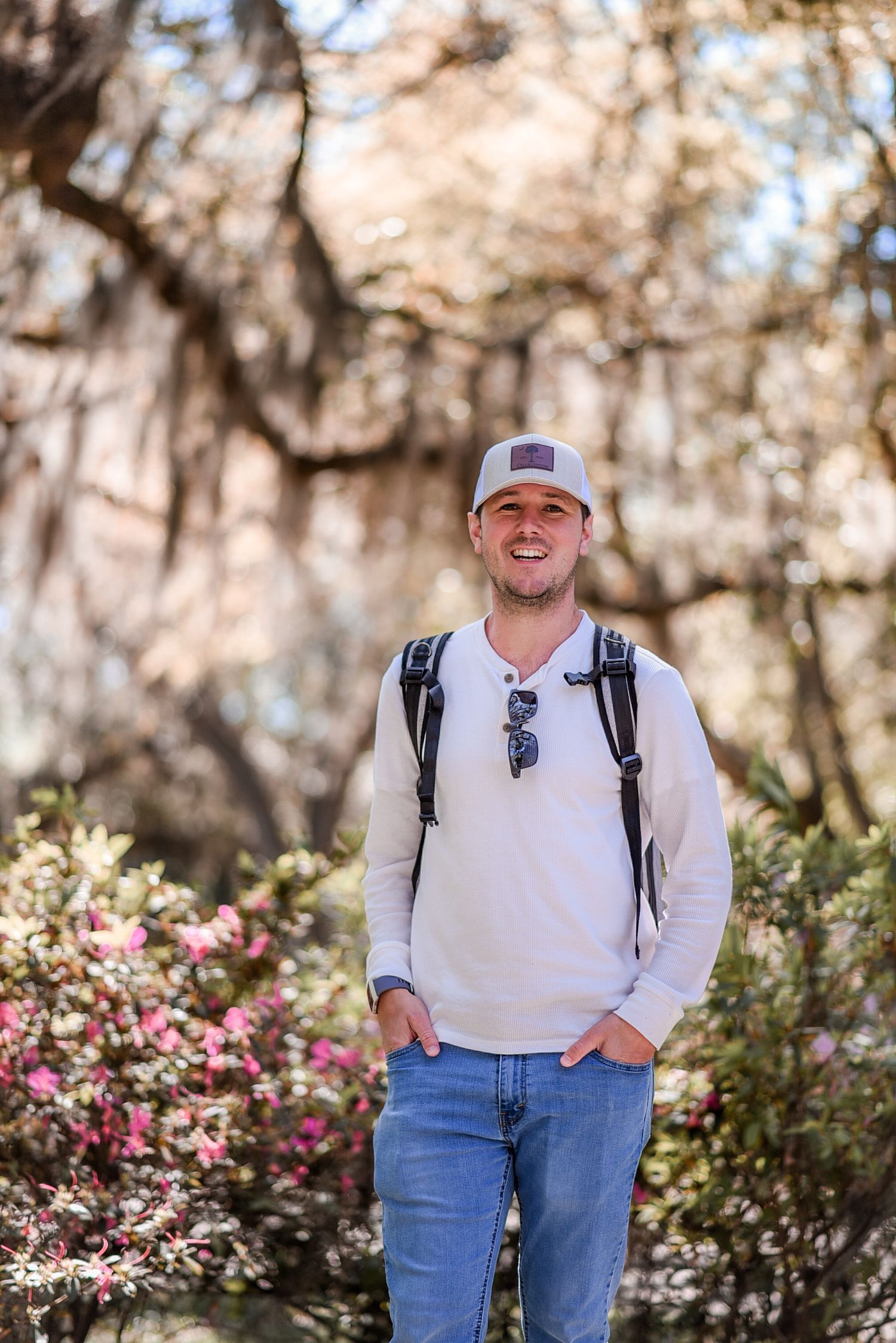 Smiling man in white long sleeve shirt and cap with backpack stands among blooming pink flowers outdoors.