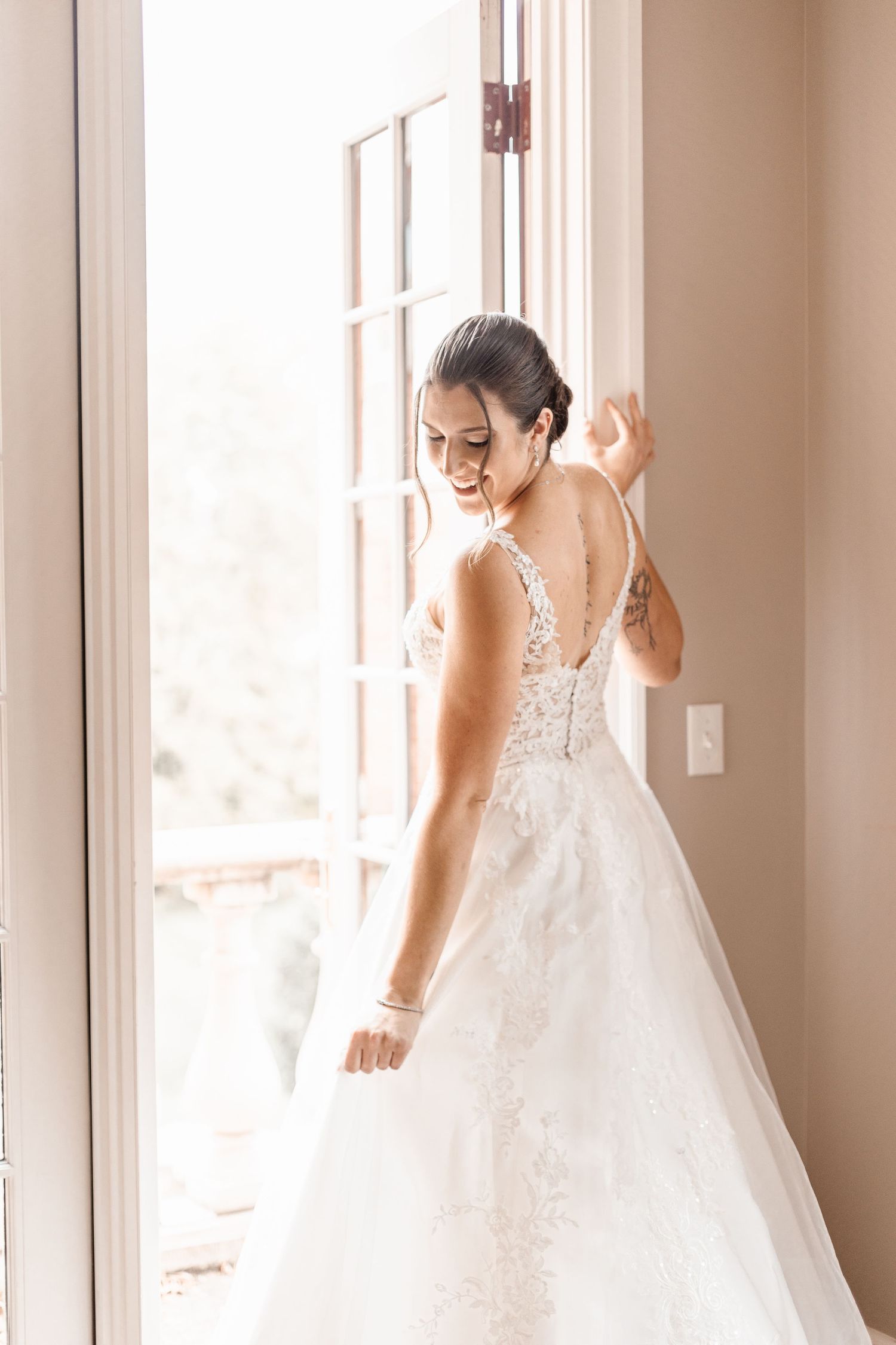Bride in a lace A-line wedding gown stands by a sunlit window adjusting her veil before the ceremony.