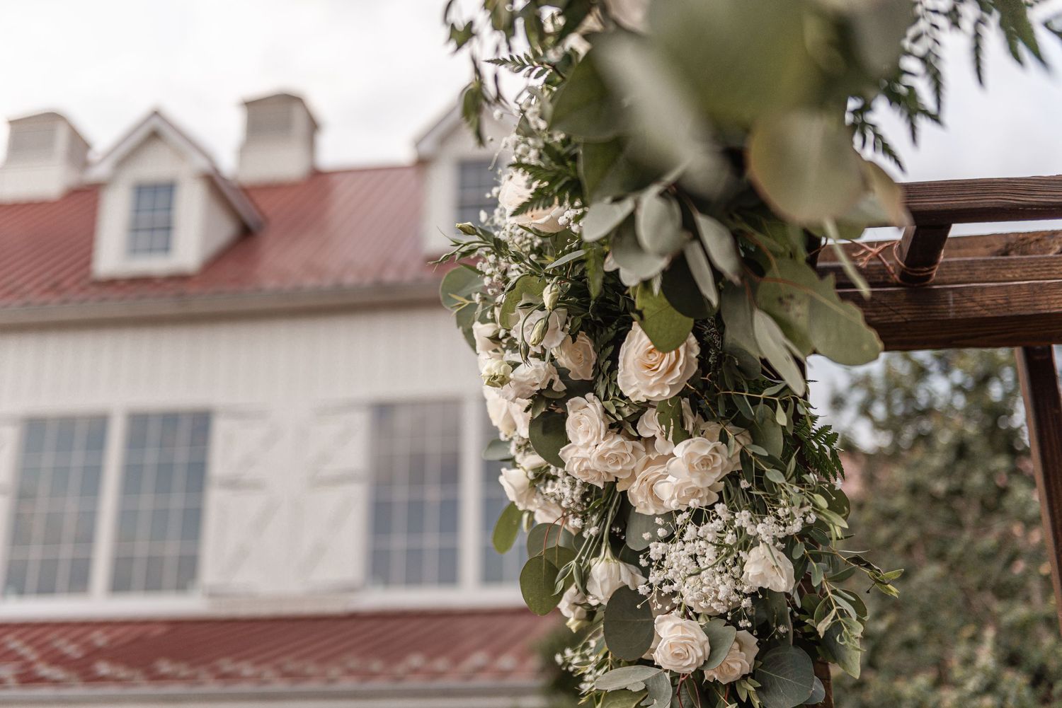 Wooden wedding arch adorned with white roses and lush greenery with a rustic venue in the background.