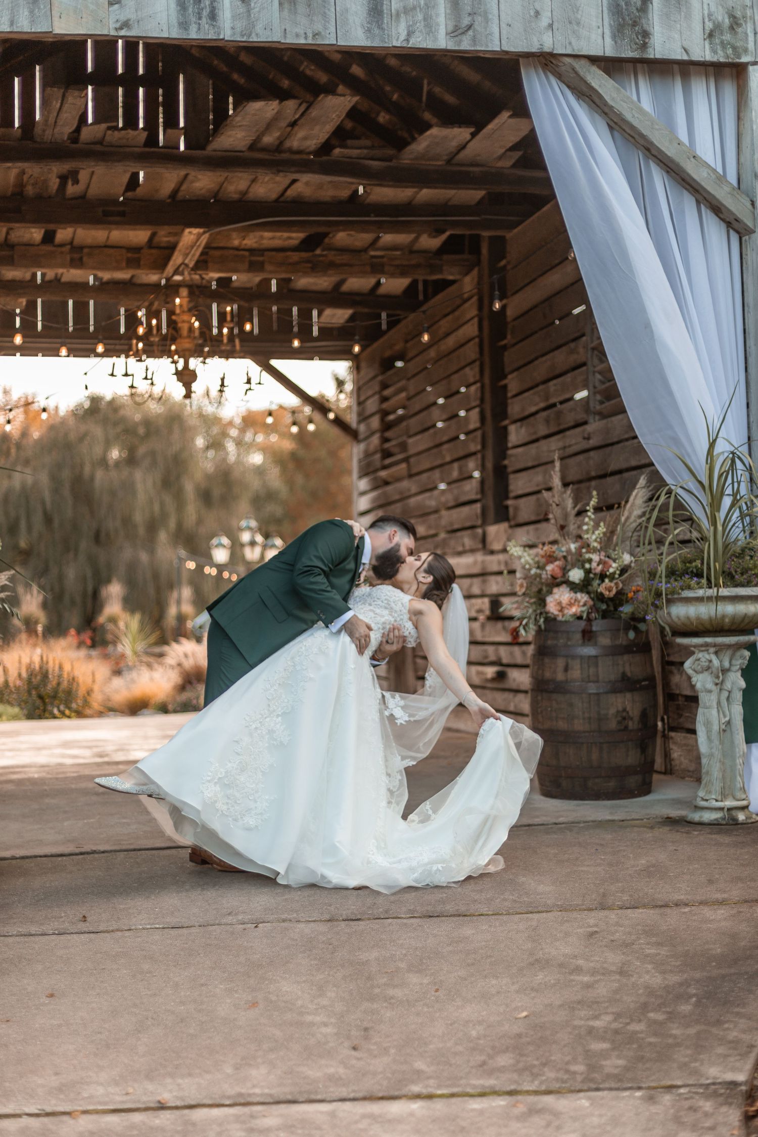 Groom dipping bride in romantic kiss under rustic wooden barn with string lights and floral decor.