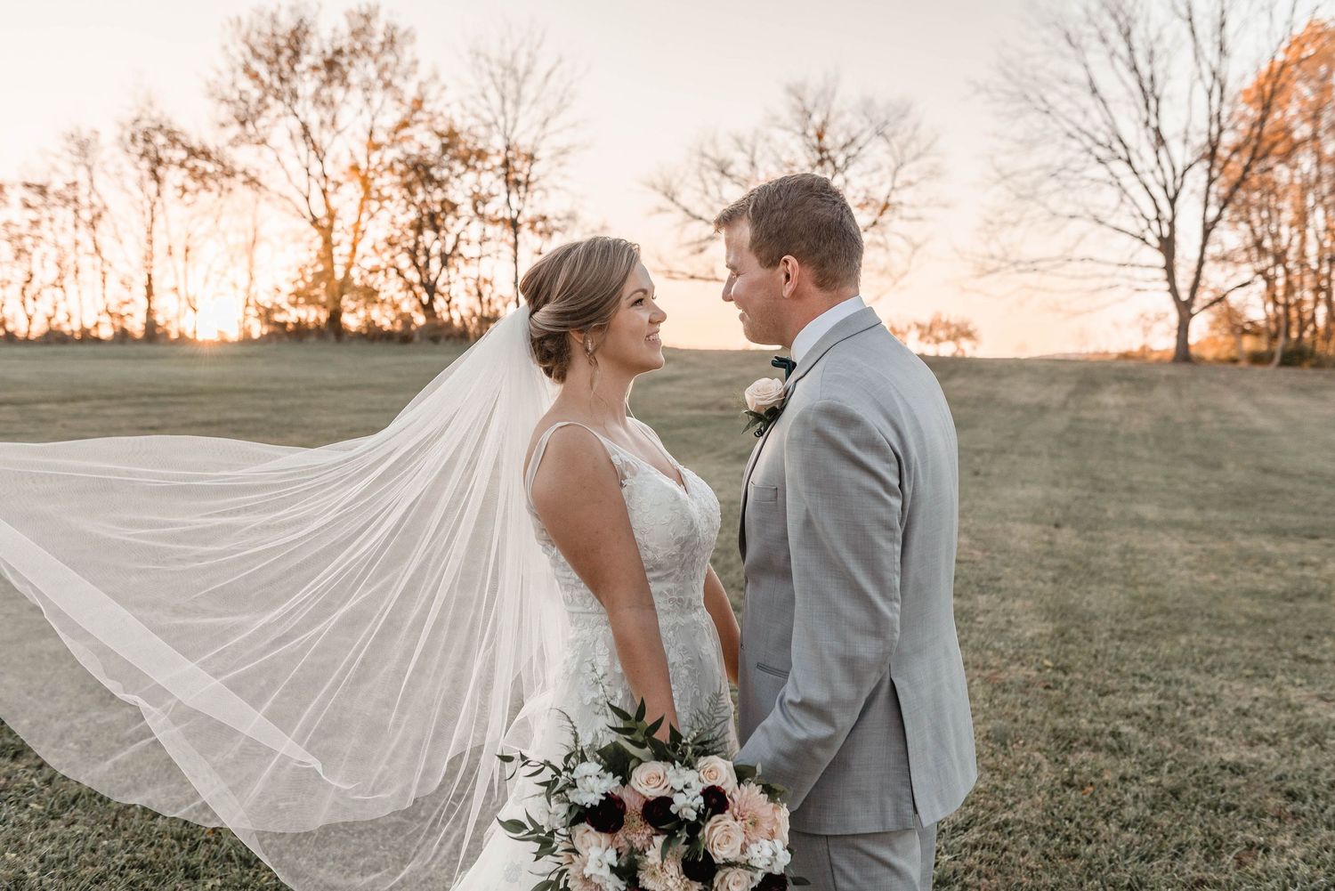 Bride and groom share a romantic moment as her veil billows at golden sunset in an open field.