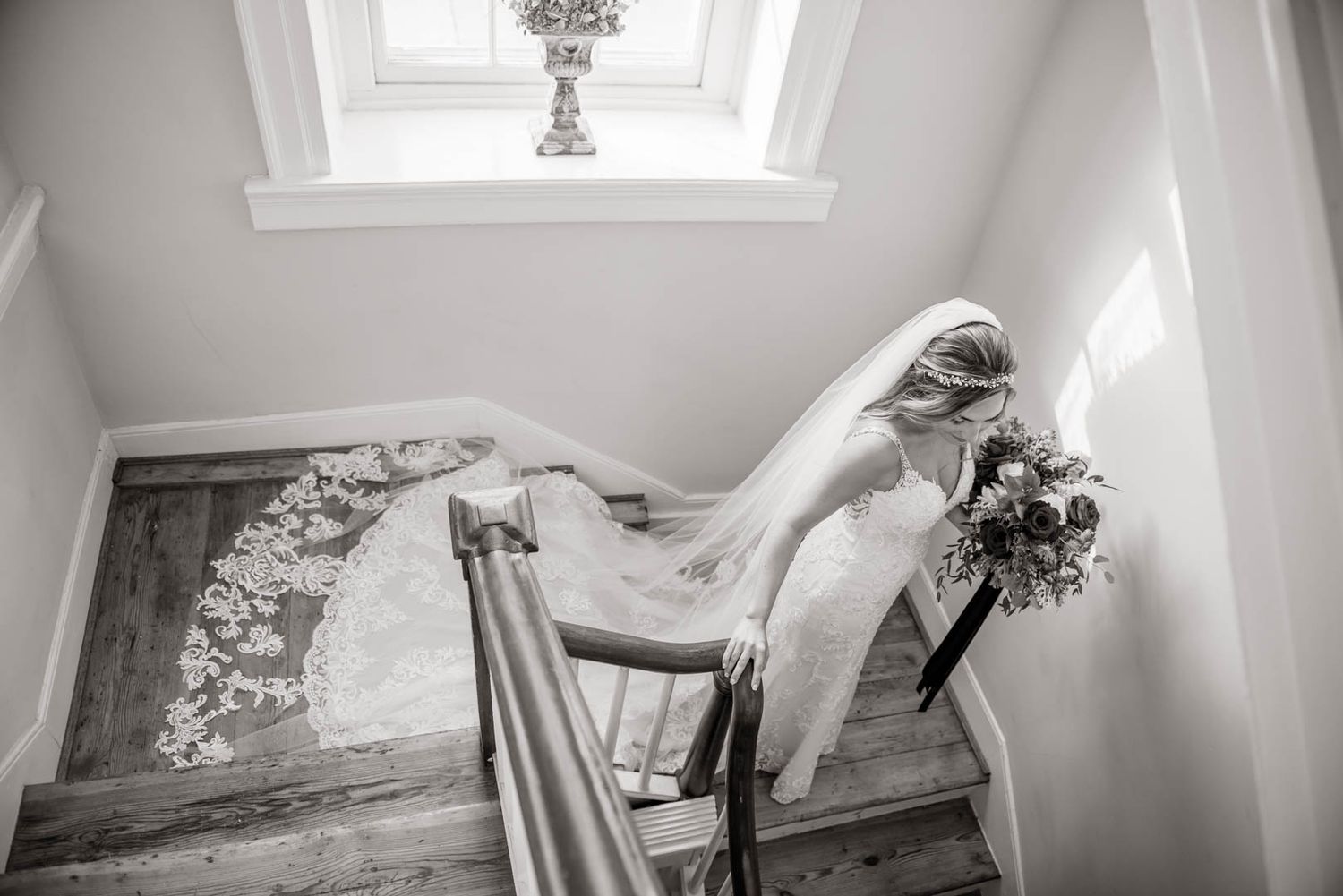 Black and white photo of a bride in a lace gown descending a sunlit staircase with a cascading veil.