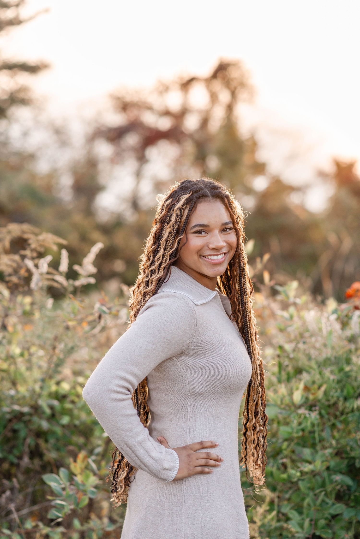 A teenage girl with long braided hair smiling confidently in a beige dress amid autumn wildflowers.