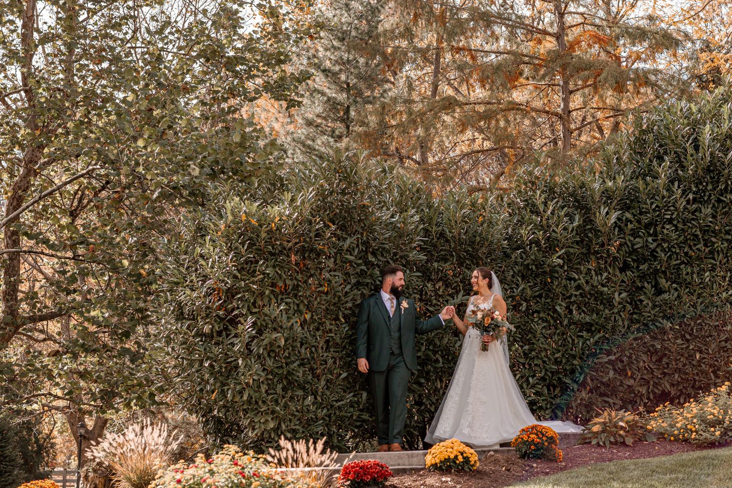 Bride and groom walking hand in hand through an autumn garden with colorful foliage and chrysanthemums.