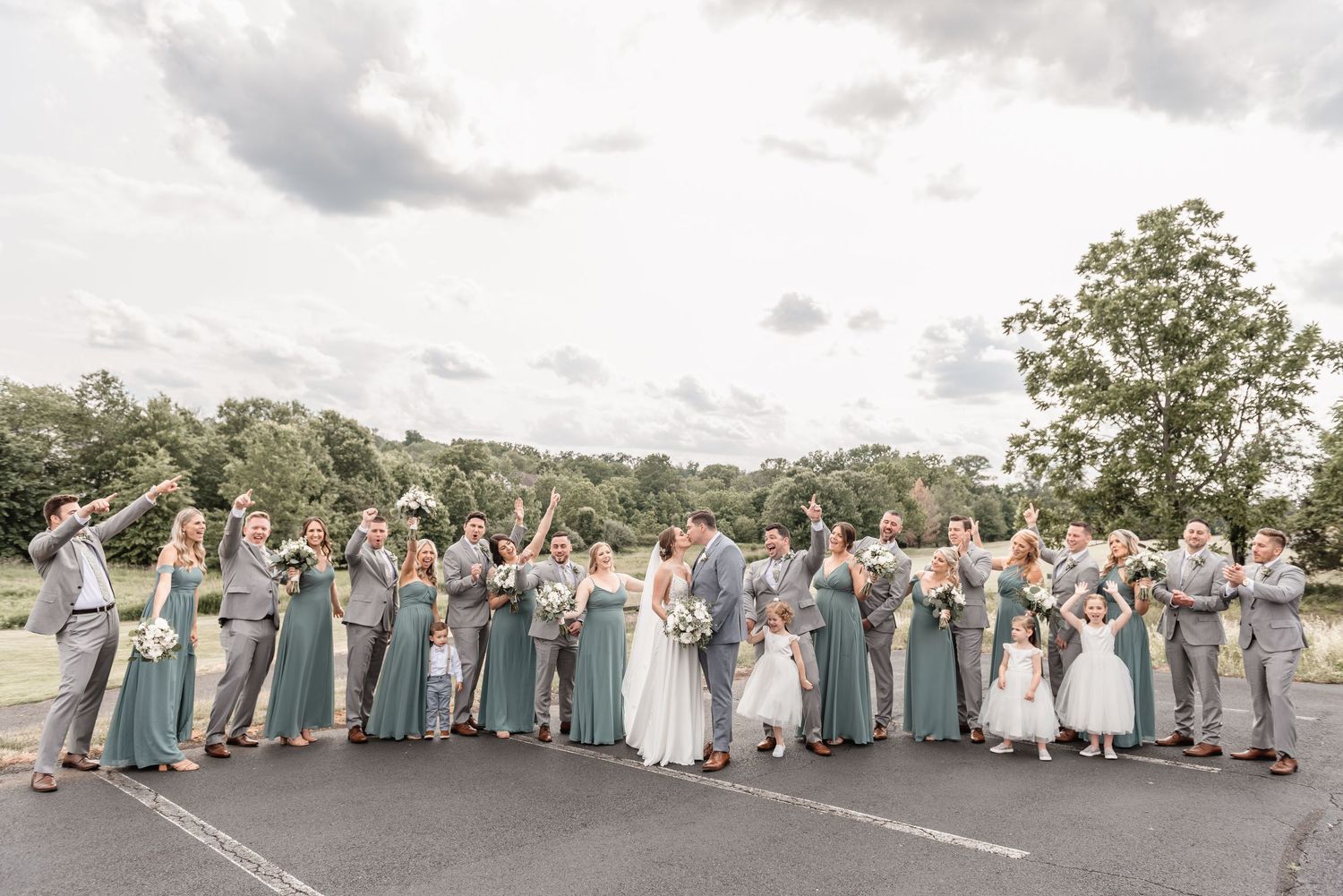 Large wedding party in sage green and gray attire posing together outdoors on a cloudy day.
