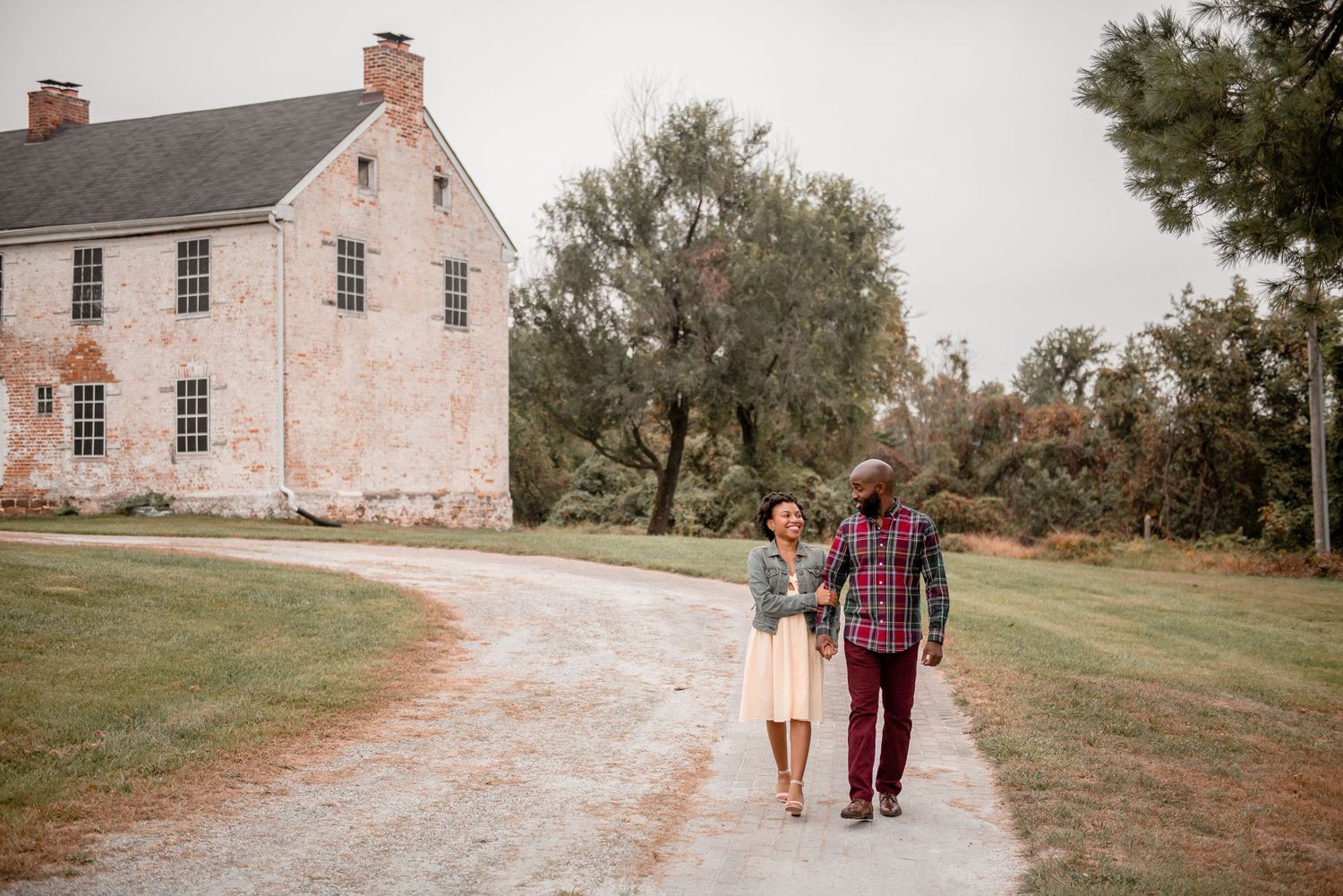 Couple walking hand in hand down gravel path toward historic brick building during fall engagement session.