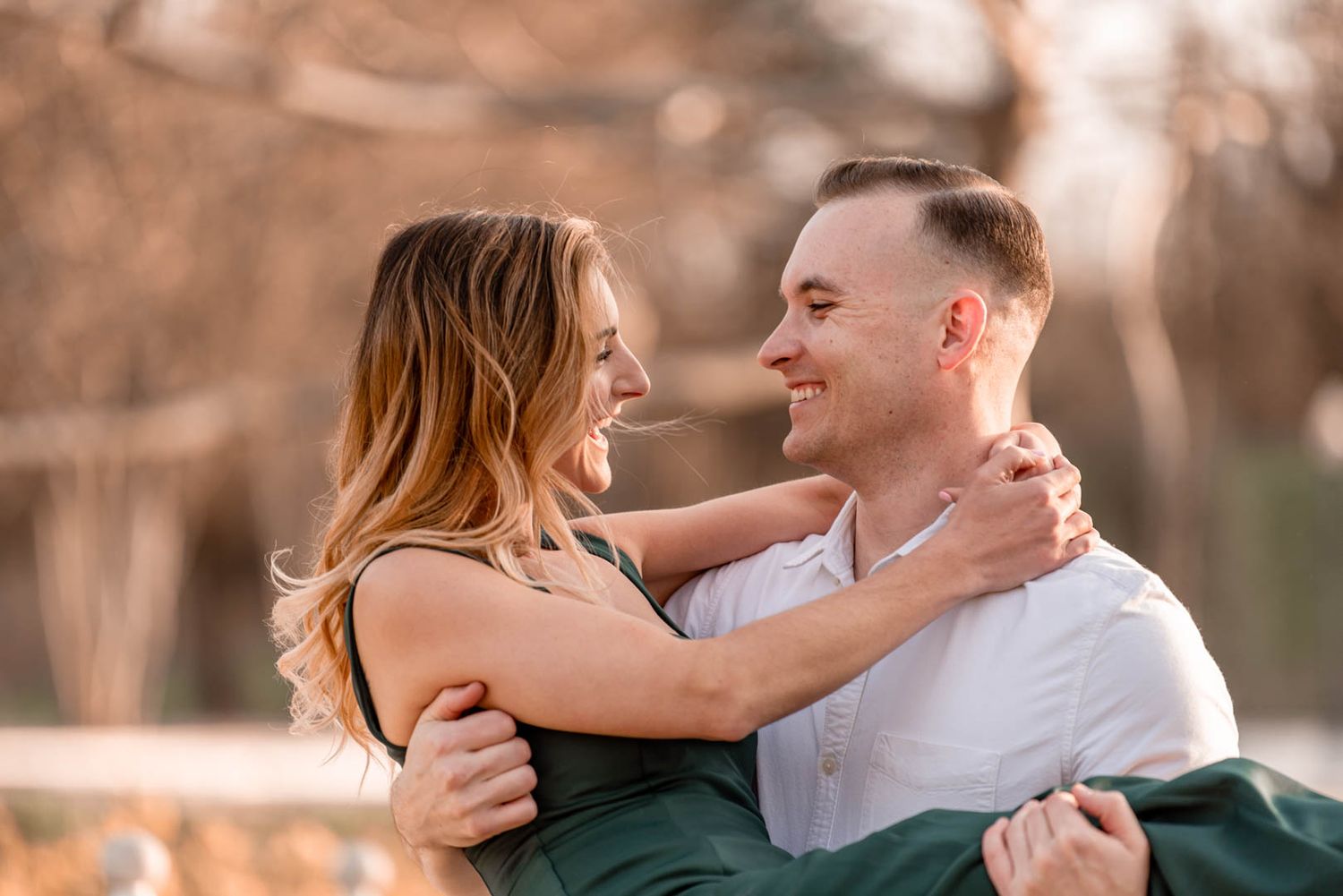 Man in white shirt lifts smiling woman in green dress during romantic outdoor engagement photo session.