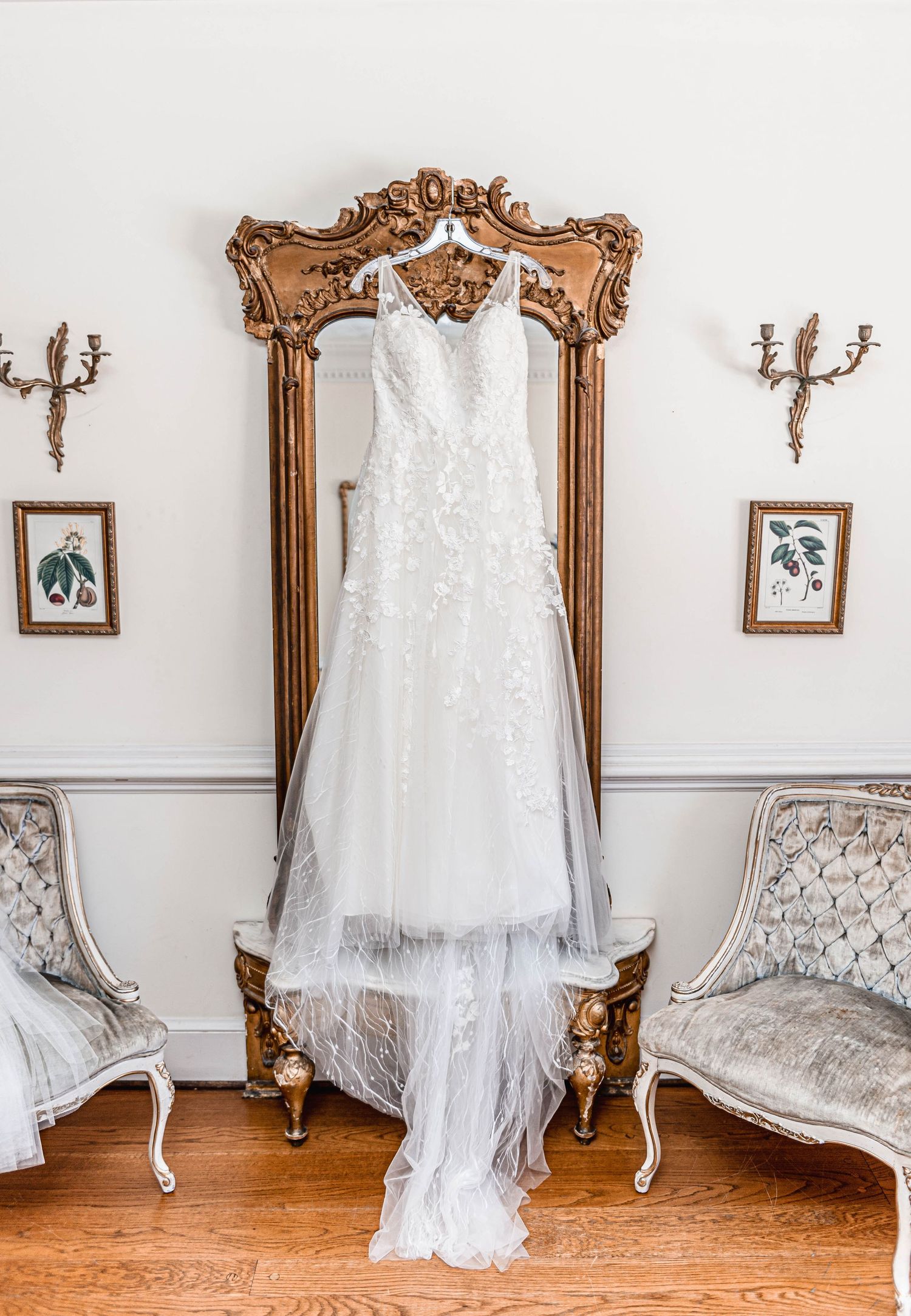 Elegant lace wedding gown hanging on an ornate gold mirror flanked by vintage chairs in a classic bridal suite.
