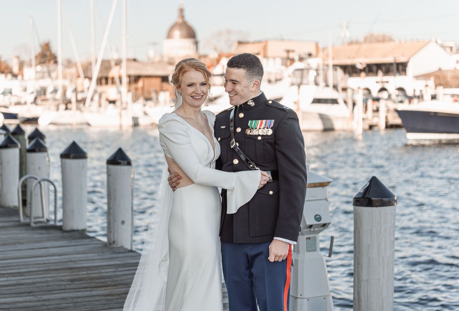 Bride and groom in Marine dress uniform pose on a waterfront dock with boats and harbor in the background.