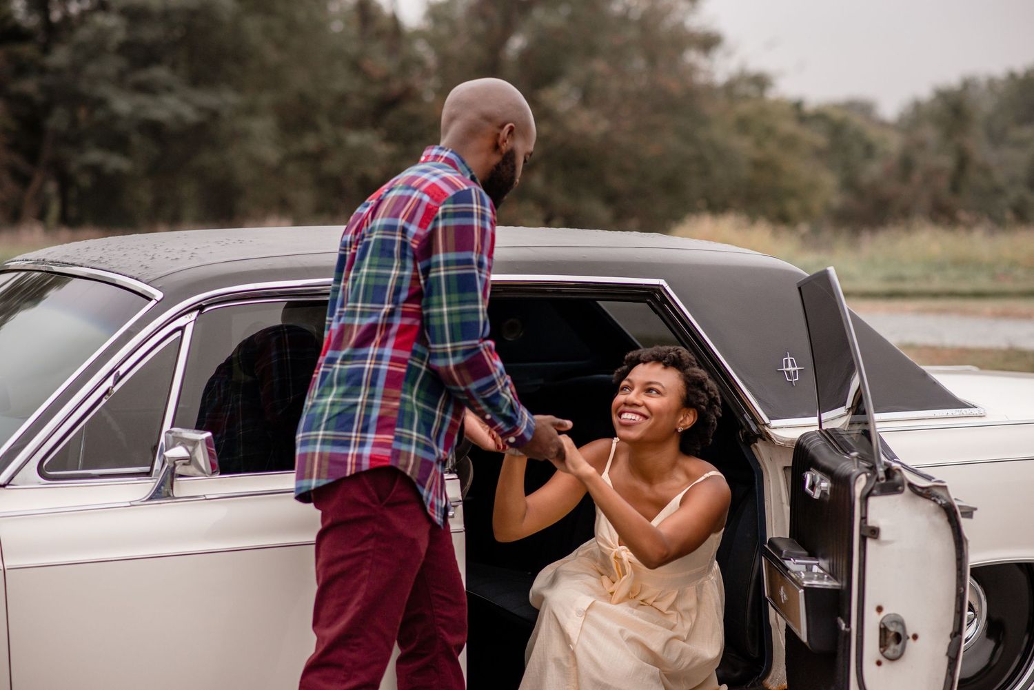 A smiling woman sits in a classic white vintage car as her partner lovingly holds her hand outside.