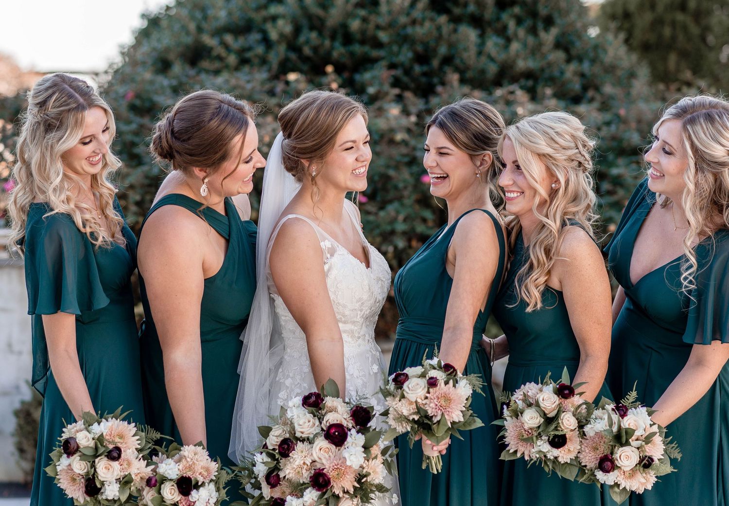 Bride surrounded by bridesmaids in dark green dresses holding blush and burgundy bouquets outdoors.