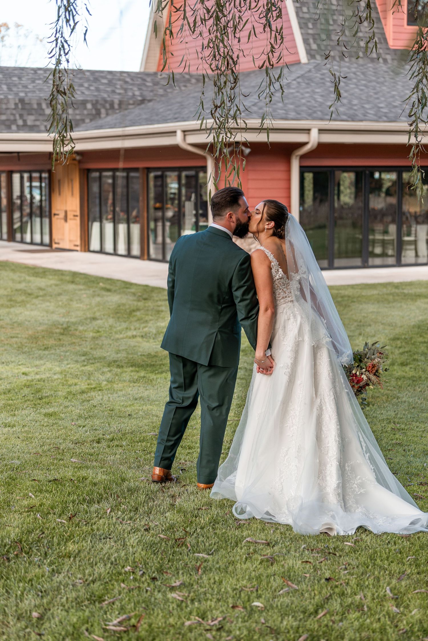Bride and groom sharing a kiss on green lawn in front of red barn venue, her lace train trailing behind.