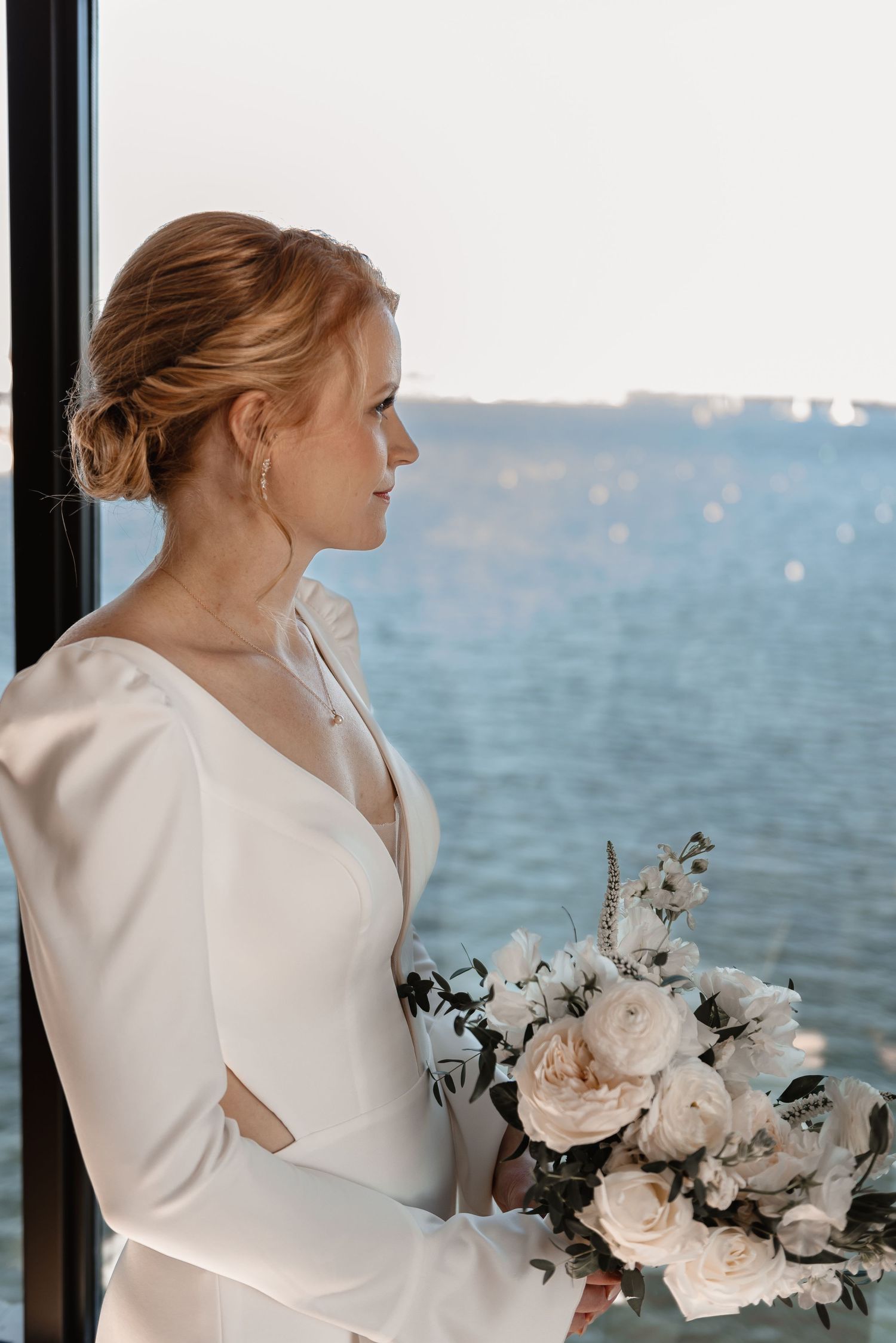 Elegant bride in a long-sleeve satin gown holding a white bouquet while gazing at a waterfront view.