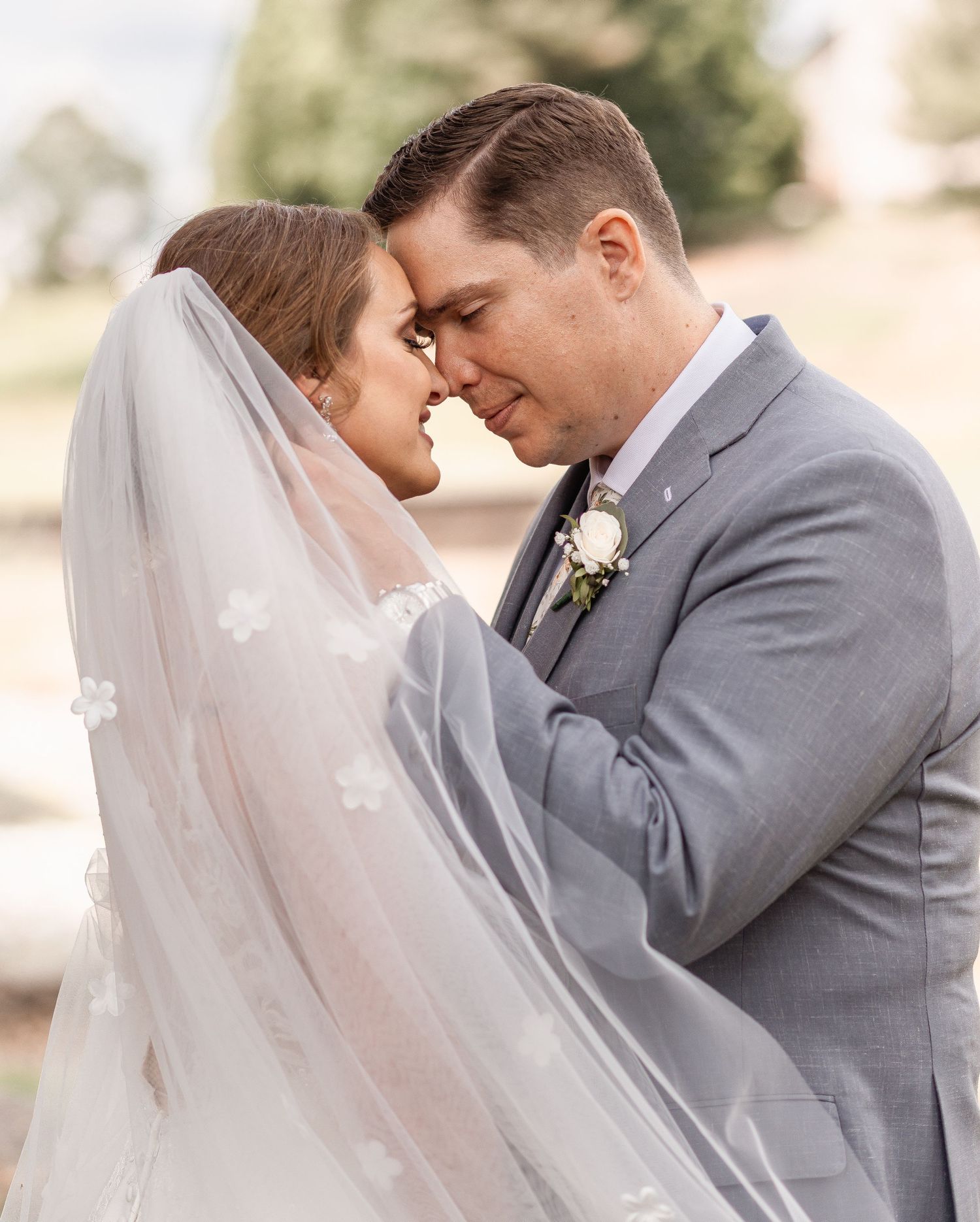 Bride and groom share an intimate forehead-to-forehead moment under a floral veil outdoors.