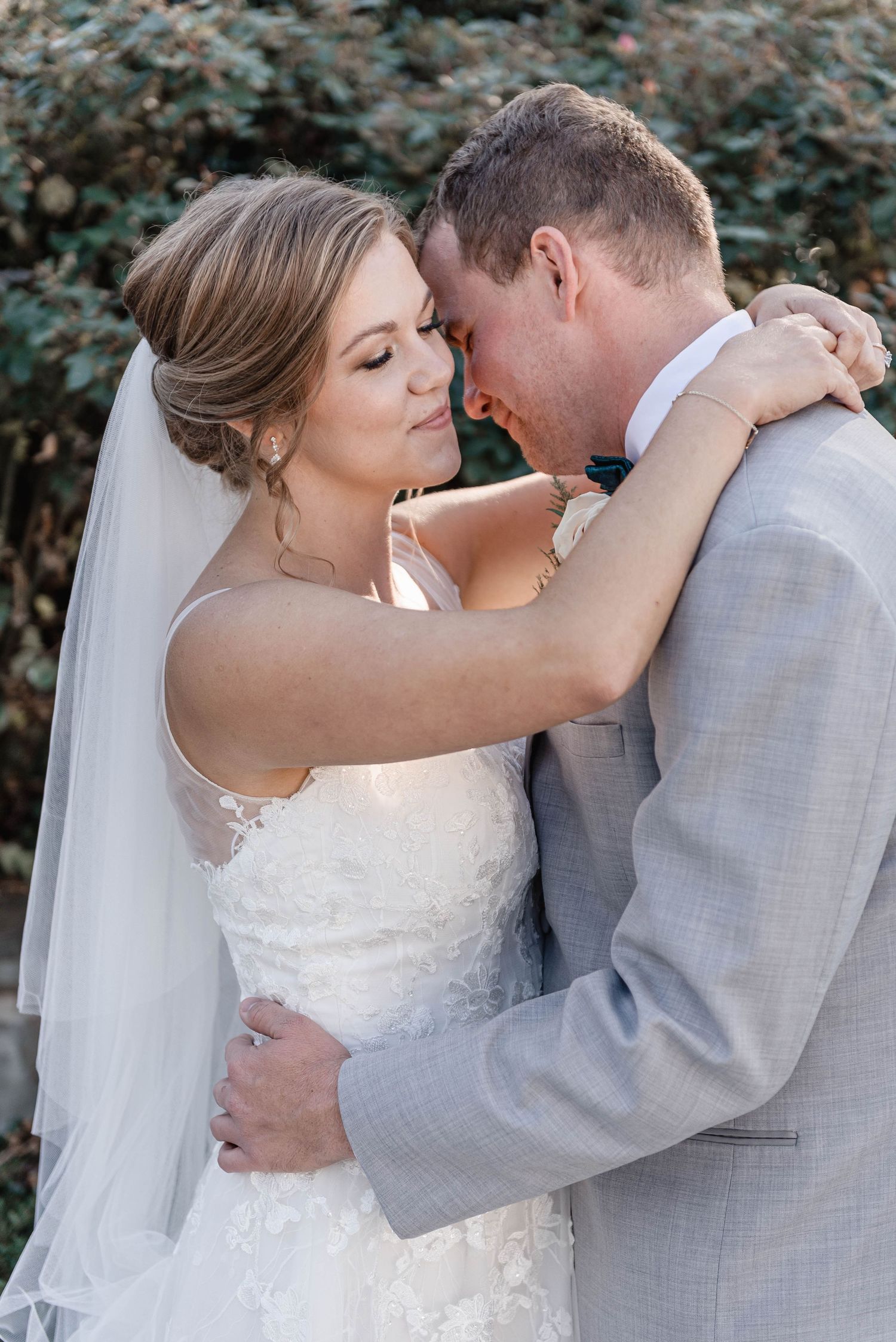 Bride in lace gown and groom in gray suit share an intimate embrace surrounded by green foliage.