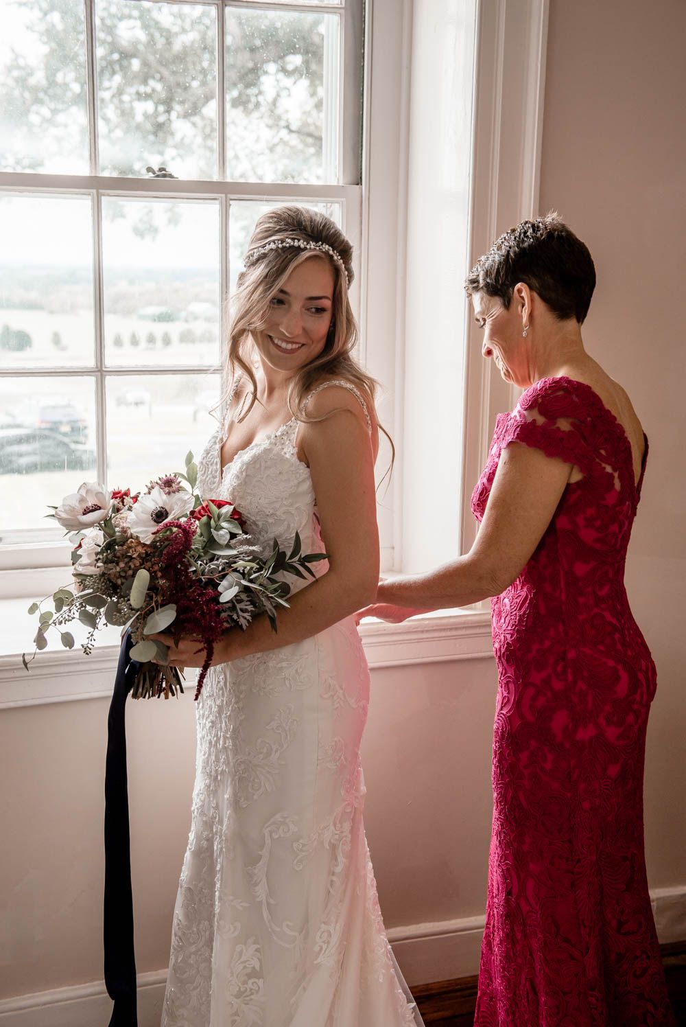 Bride in lace wedding gown holds bouquet while sharing a joyful moment with mother in red dress.