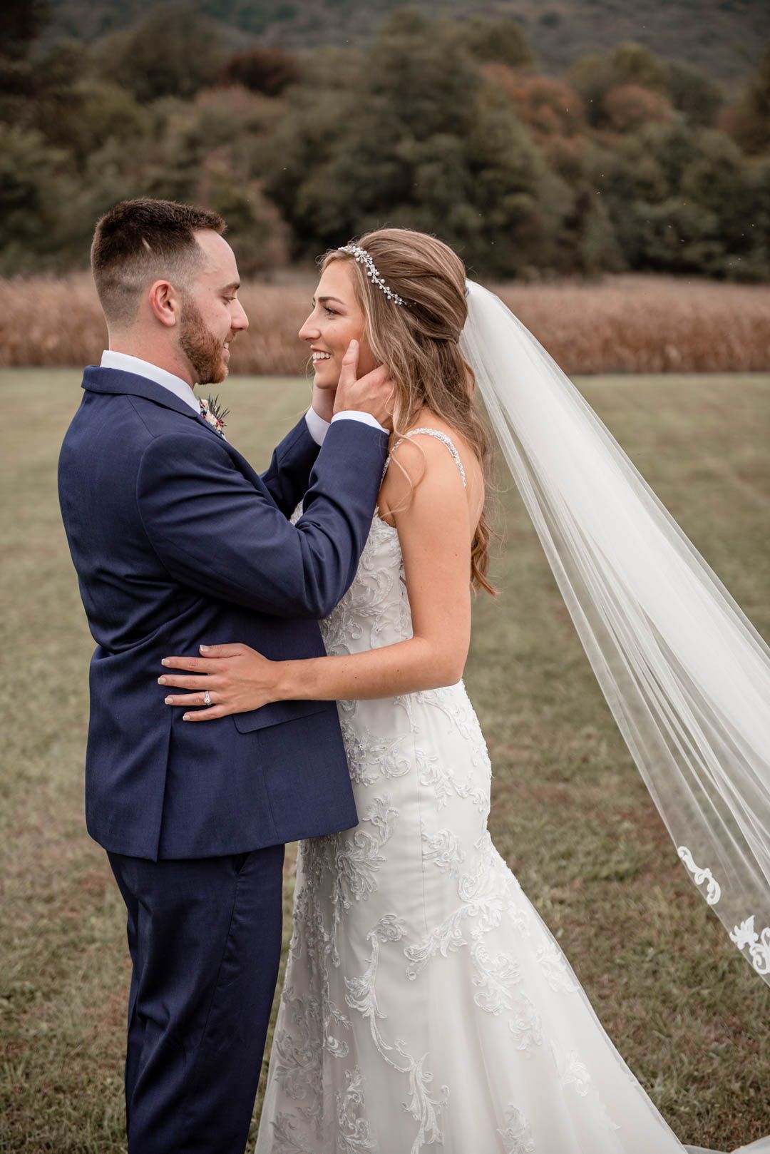 Groom tenderly cups brides face as she smiles joyfully in a lace wedding gown in a scenic outdoor field.
