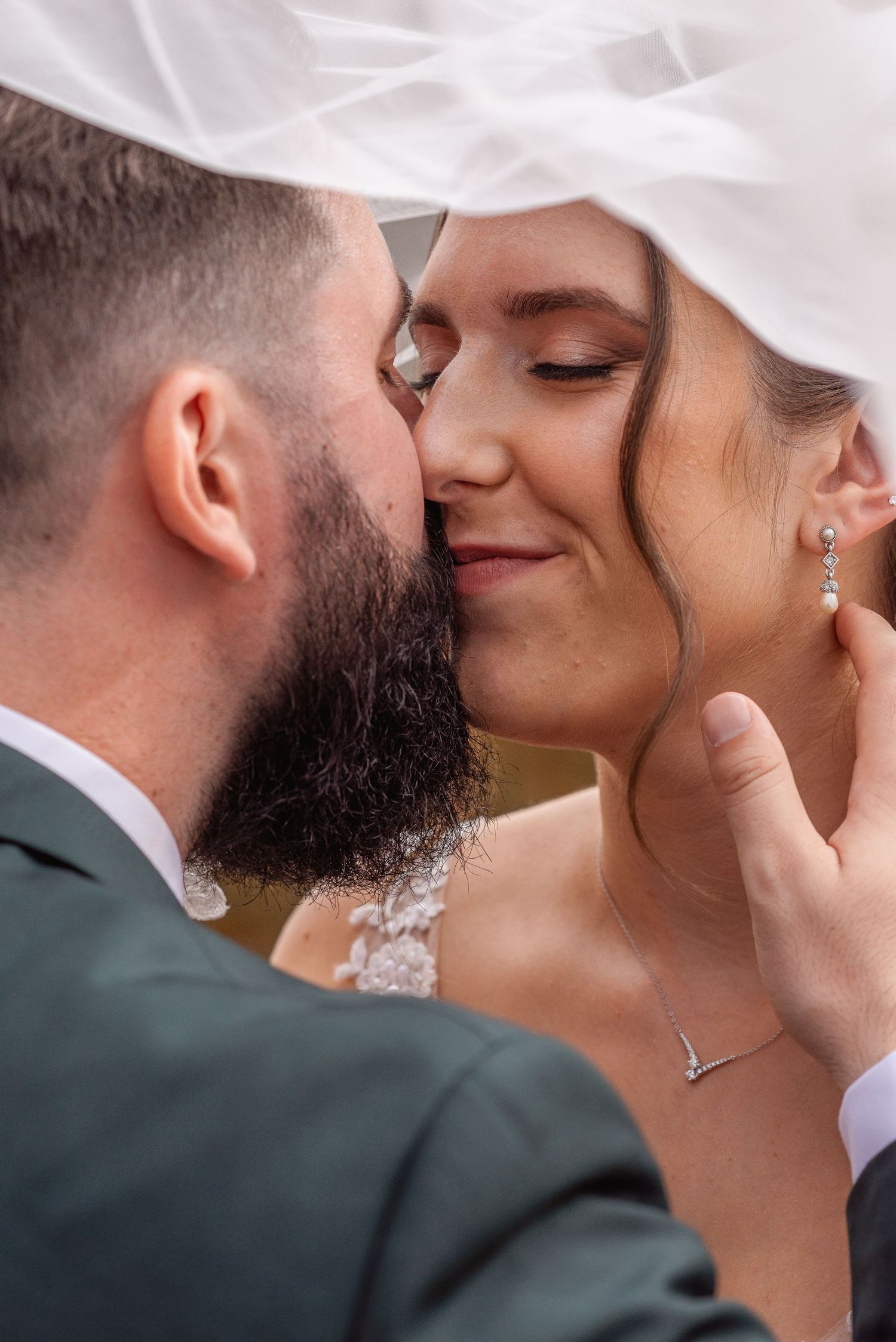 Groom tenderly kissing bride on cheek under her veil, bride smiling with eyes closed wearing pearl earrings.