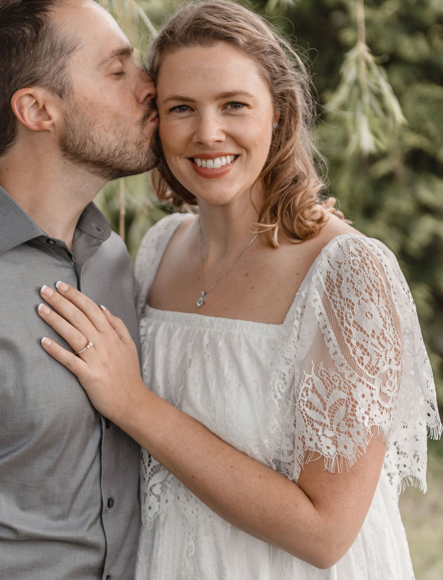 Man kissing woman on cheek as she smiles wearing a white lace dress with an engagement ring.