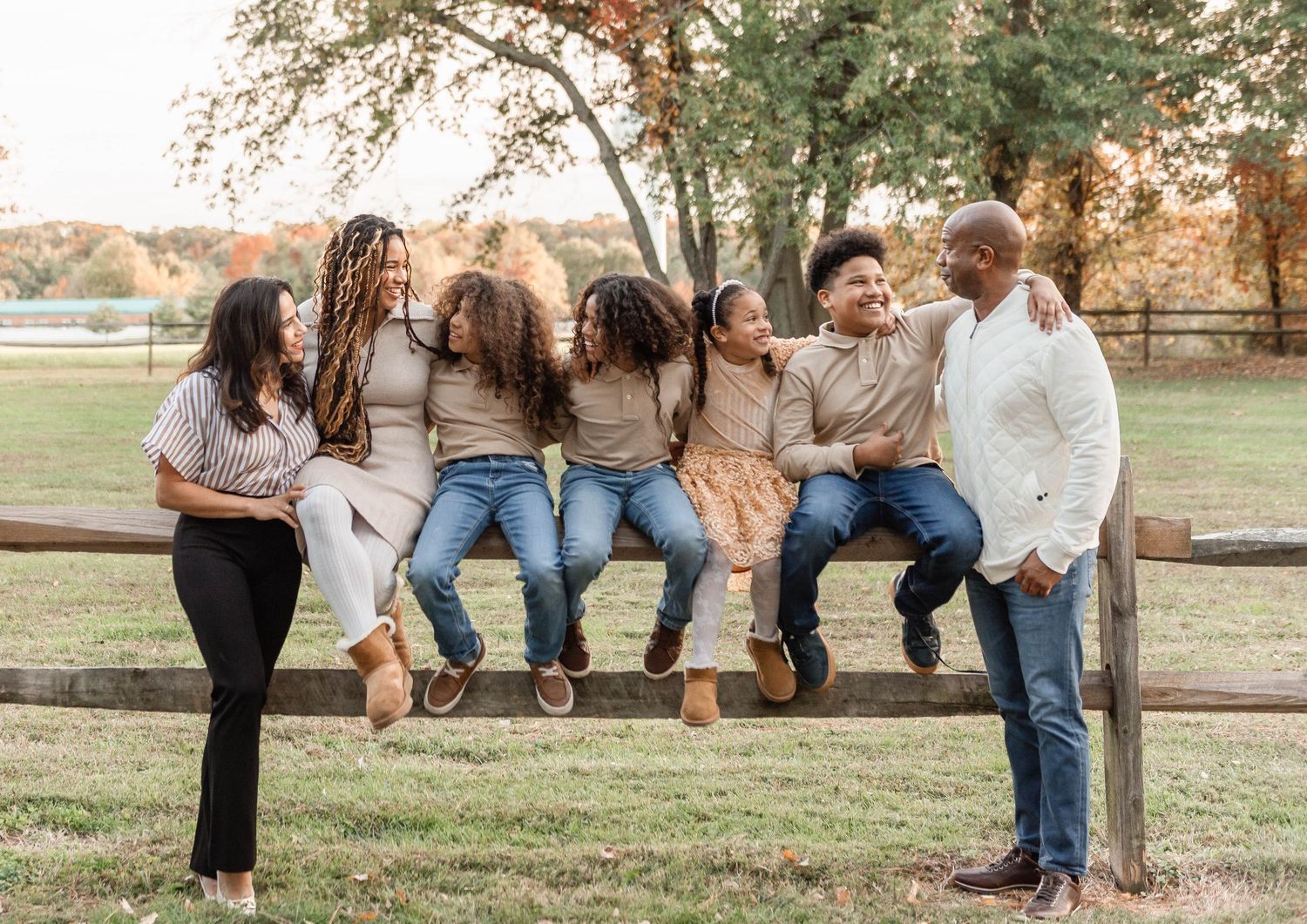 A cheerful family of six sitting and laughing together on a wooden fence in an open autumn field.