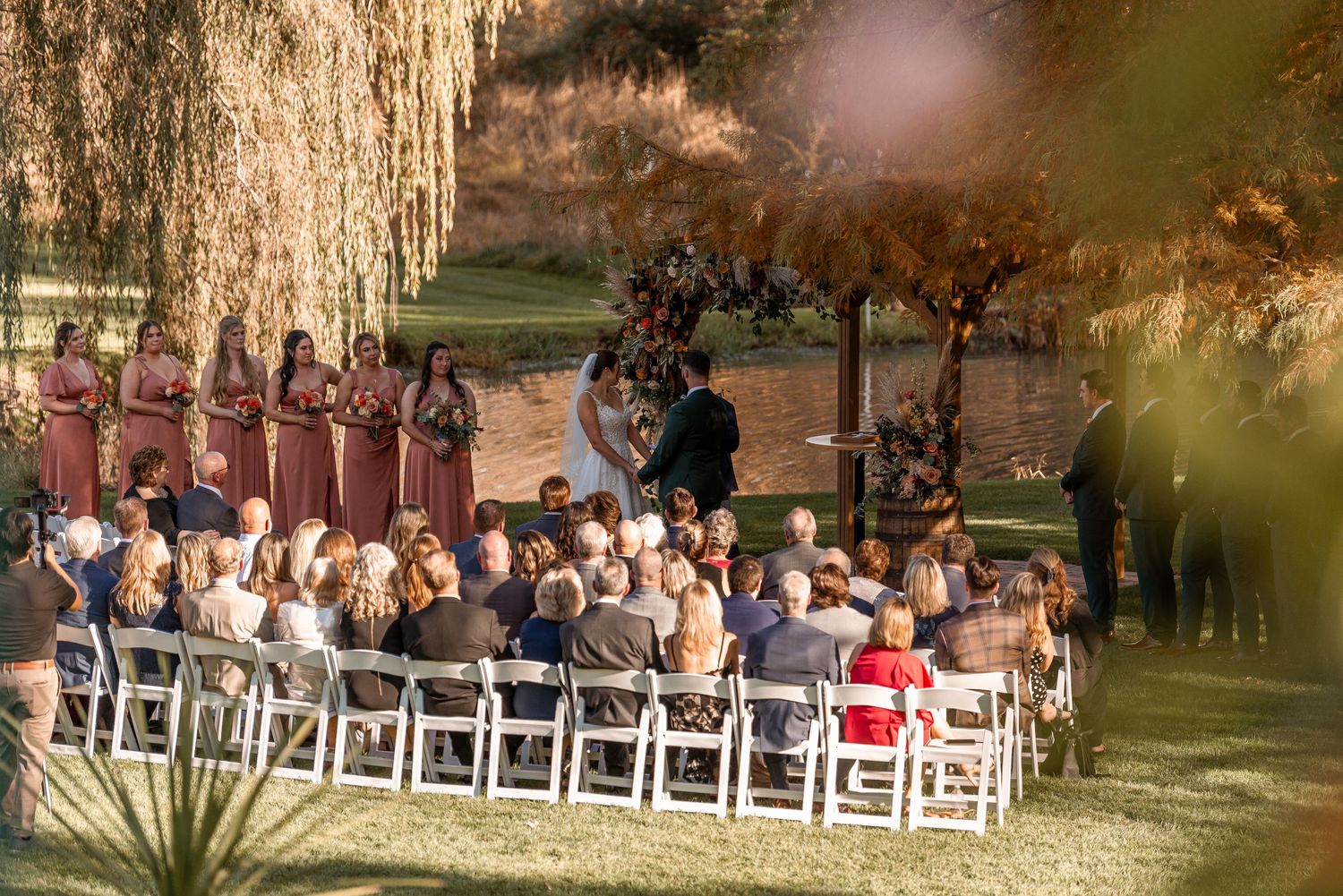 Outdoor fall wedding ceremony with guests seated on white chairs beneath weeping willow trees.