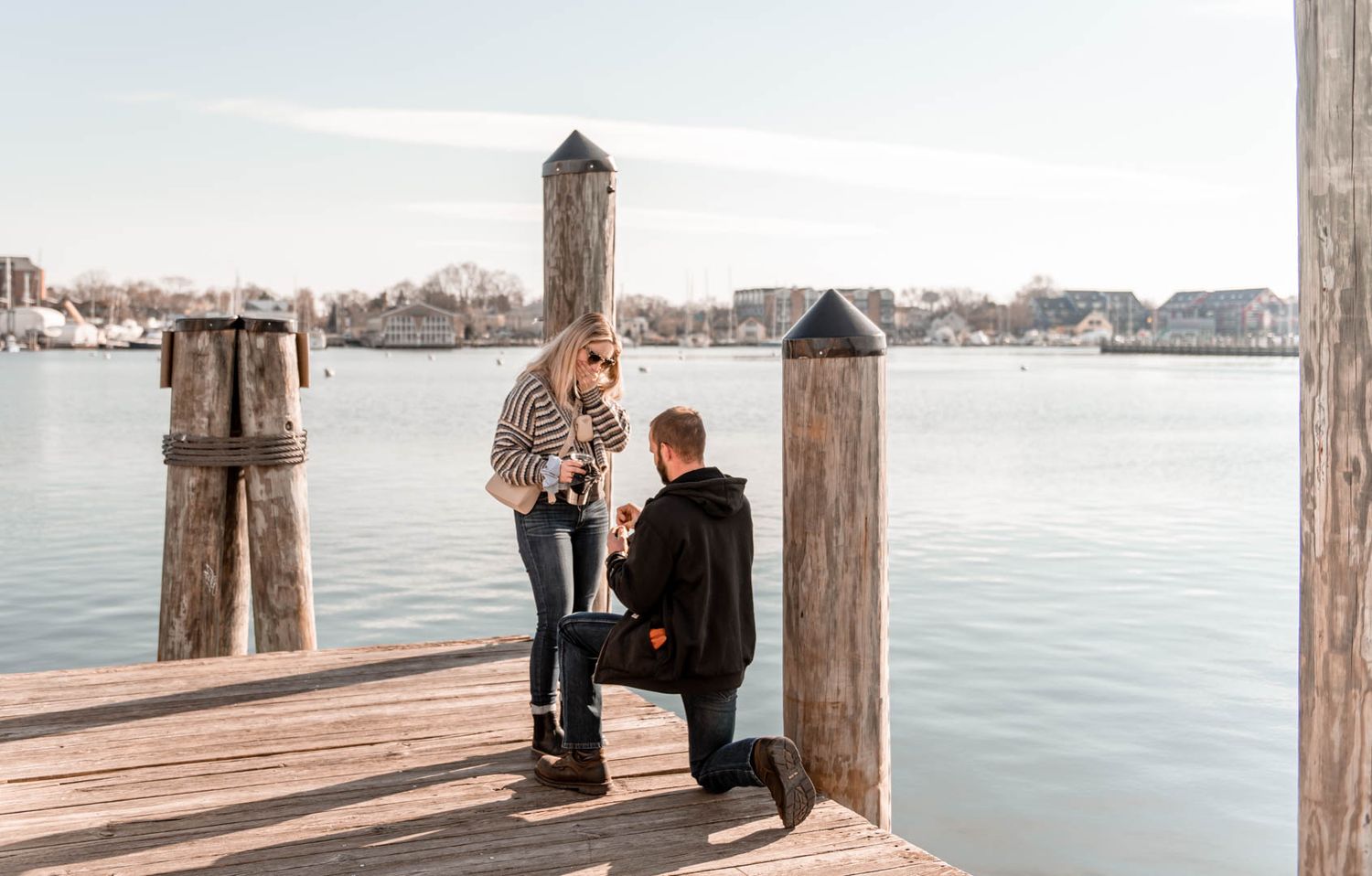Man proposing on one knee to woman on a waterfront dock on a sunny winter day.