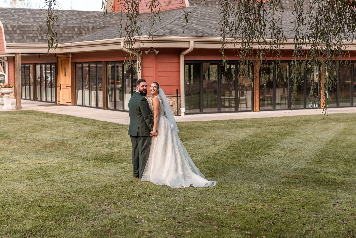 Bride and groom standing on green lawn in front of red barn wedding venue under a weeping willow tree.