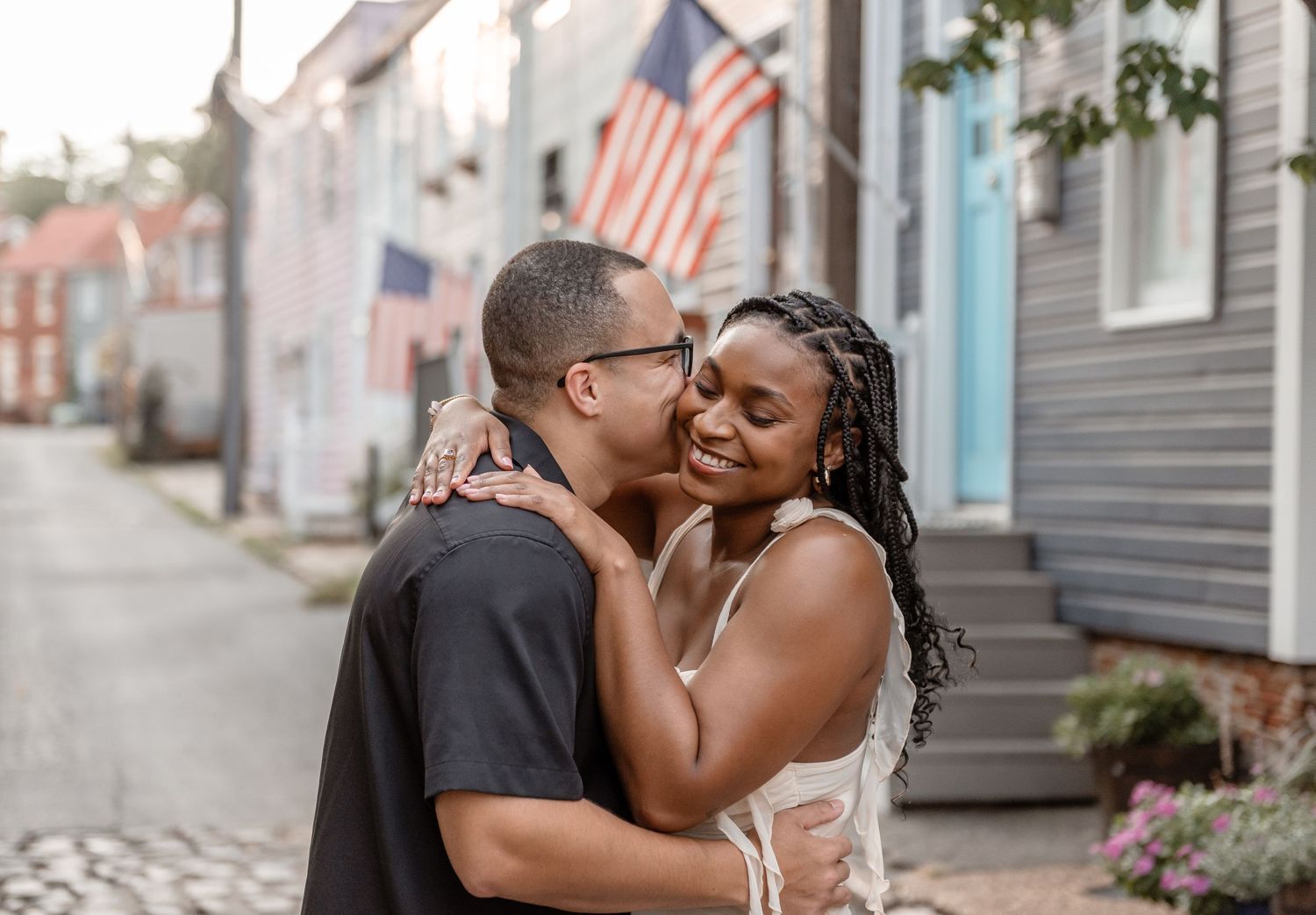 Romantic couple sharing a tender moment on a cobblestone street with an American flag backdrop.