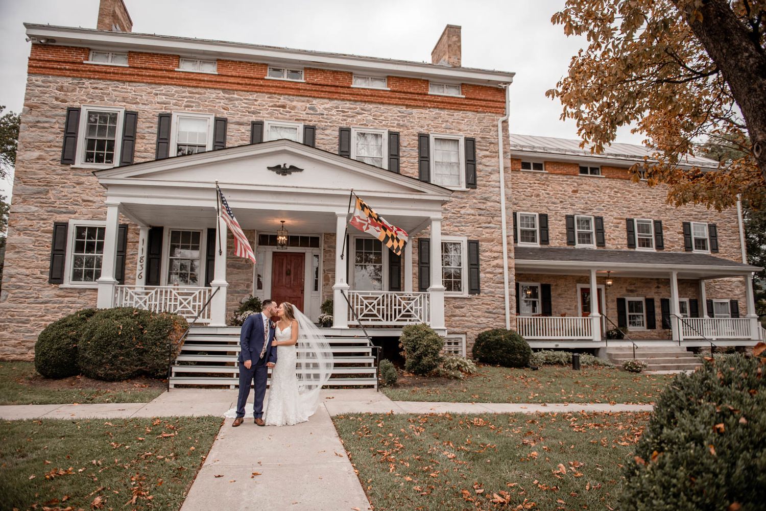 Bride and groom share a kiss in front of a historic brick colonial wedding venue with American flags.
