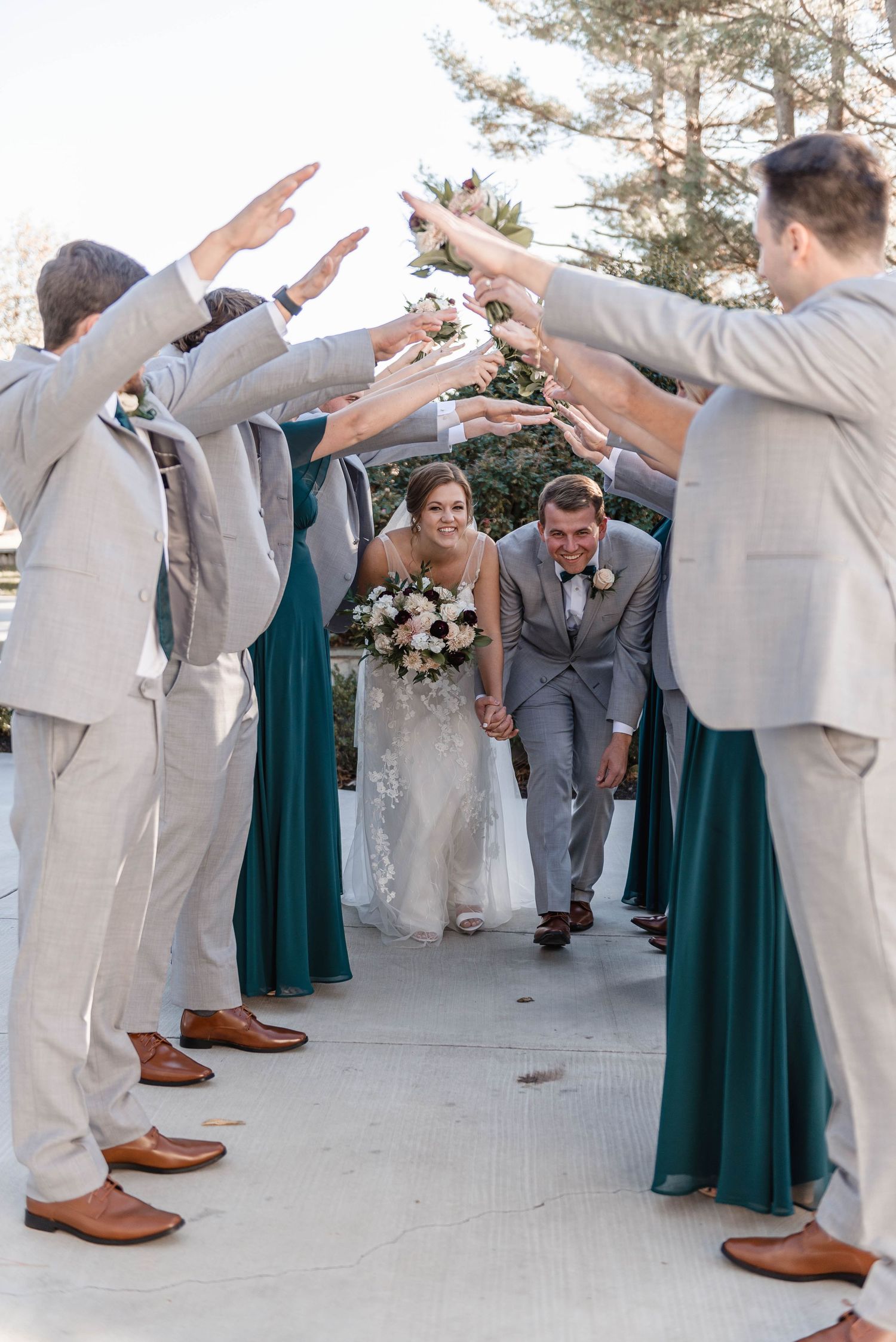 Newlyweds walk through a celebratory arch formed by groomsmen and bridesmaids in grey and emerald attire.
