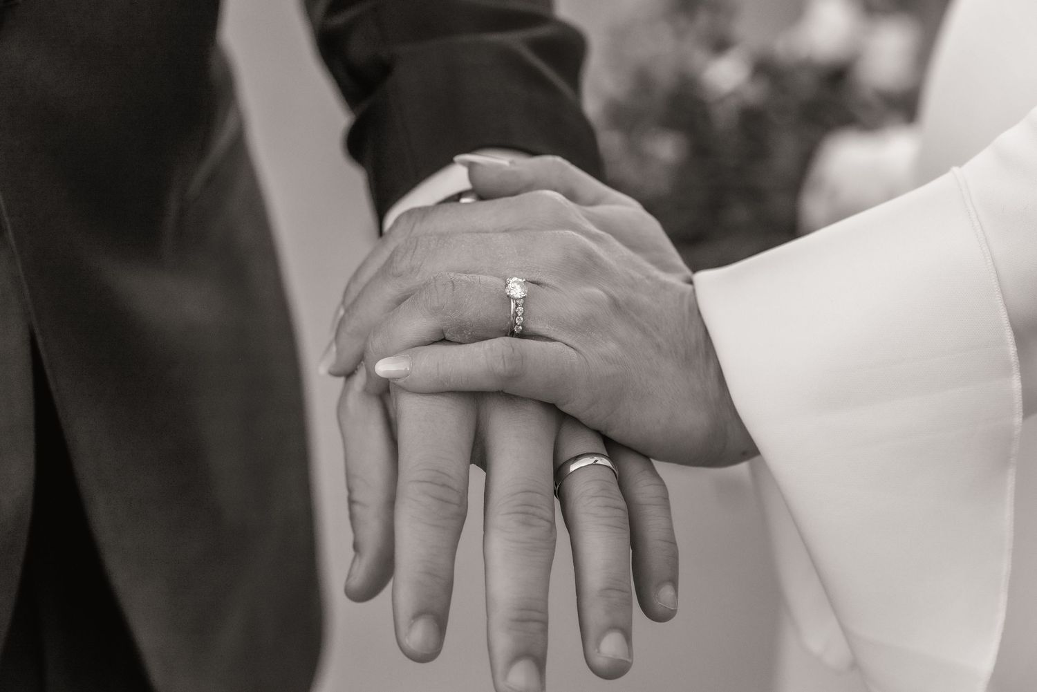 Black and white close-up of newlyweds hands showing diamond engagement ring and wedding bands.